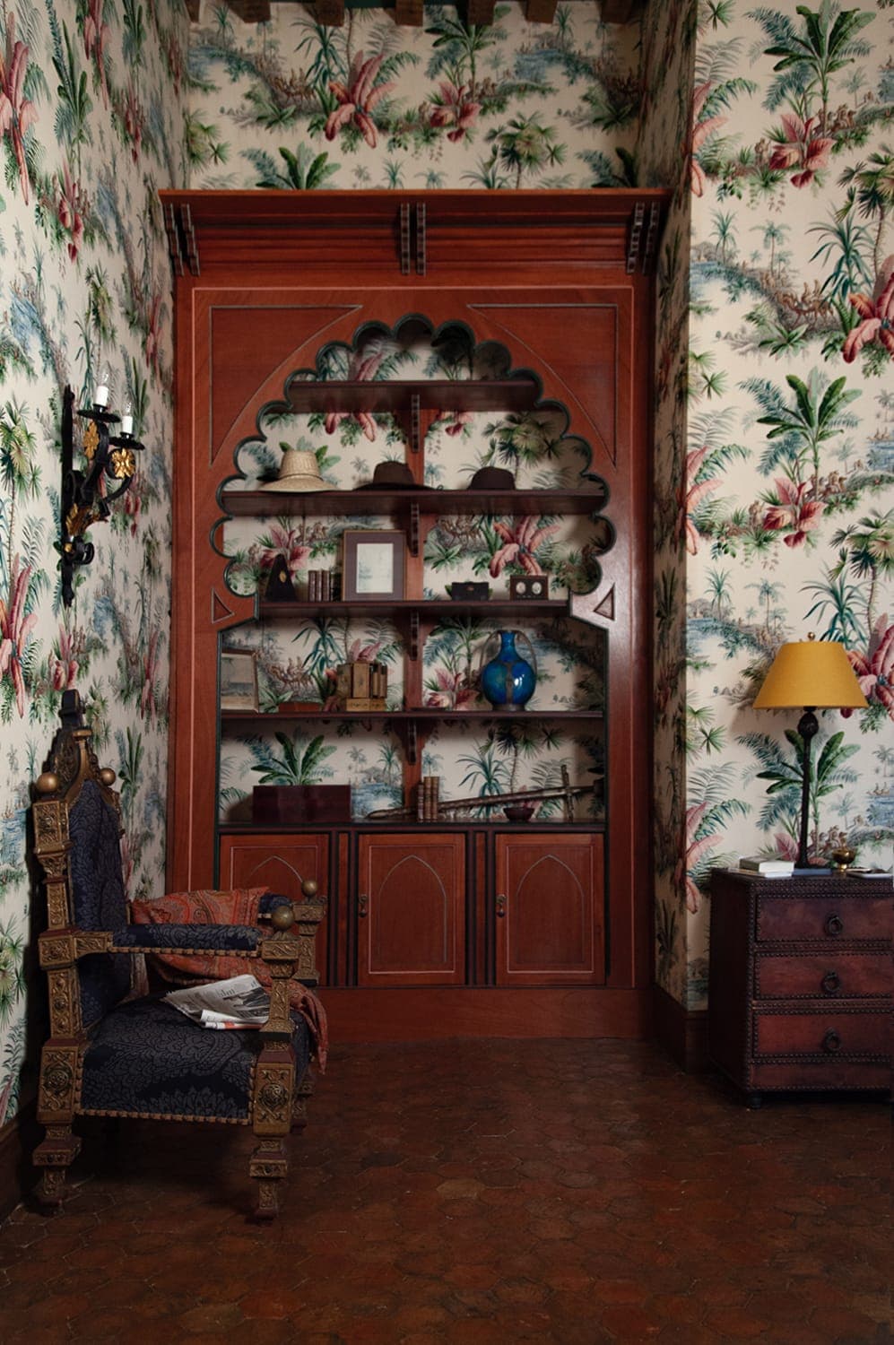 A cosy room with a wooden bookcase set in a decorative alcove. The walls are adorned with floral wallpaper. An ornate armchair and a wooden bedside table with a lamp add to the vintage decor. A few objects are neatly arranged on the shelves.