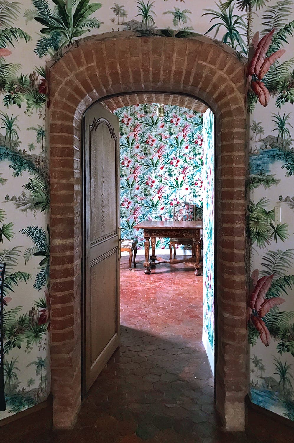 View through a vaulted brick door into a room with vibrant tropical wallpaper depicting palm trees and flowers. The floor is of terracotta tiles and there is a wooden table and chairs in the room.