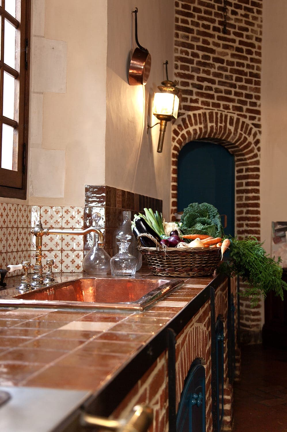 A rustic kitchen with red brick walls and tiled countertops. The copper sink and faucet are visible. A basket filled with fresh vegetables, including carrots and kale, sits on the counter. Vintage-style light fixtures adorn the walls.