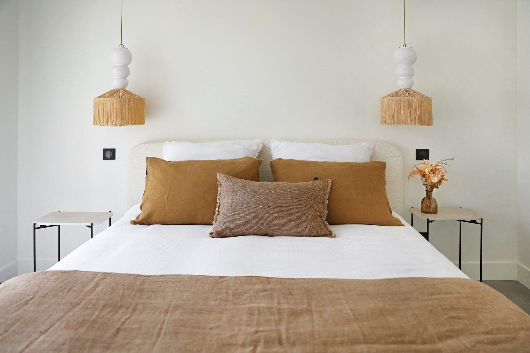 A comfortable bedroom with a well-made bed features brown and white bedding. Two fringed hanging lamps are suspended on either side above small side tables. A vase with dried flowers sits on the table to the right against a light wall.