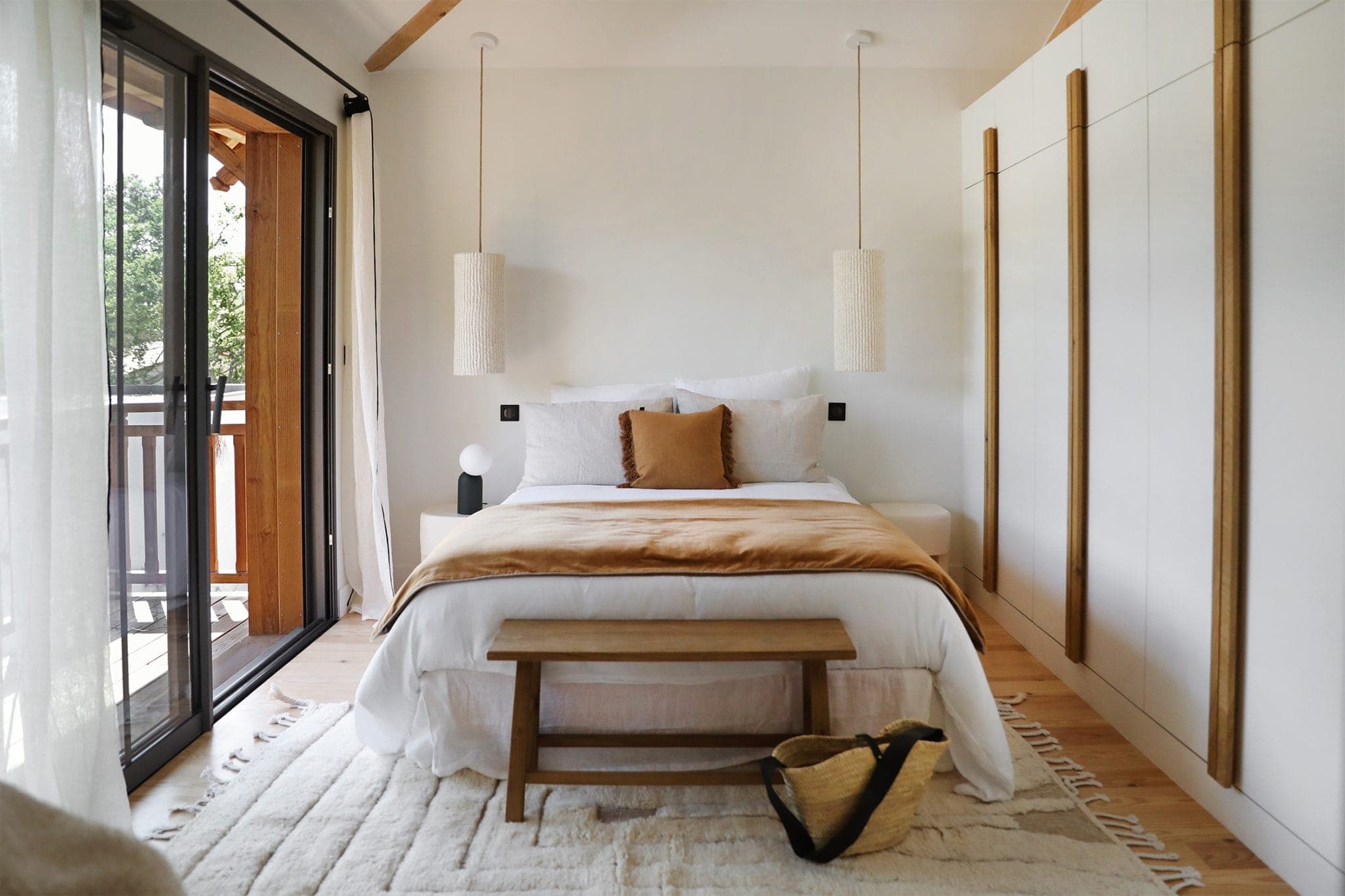 A comfortable room with a king-size bed with white and beige pillows and a brown blanket. Hanging lights hang from the ceiling and large glass doors open onto a balcony. A small wooden bench and a woven basket lie on the floor.