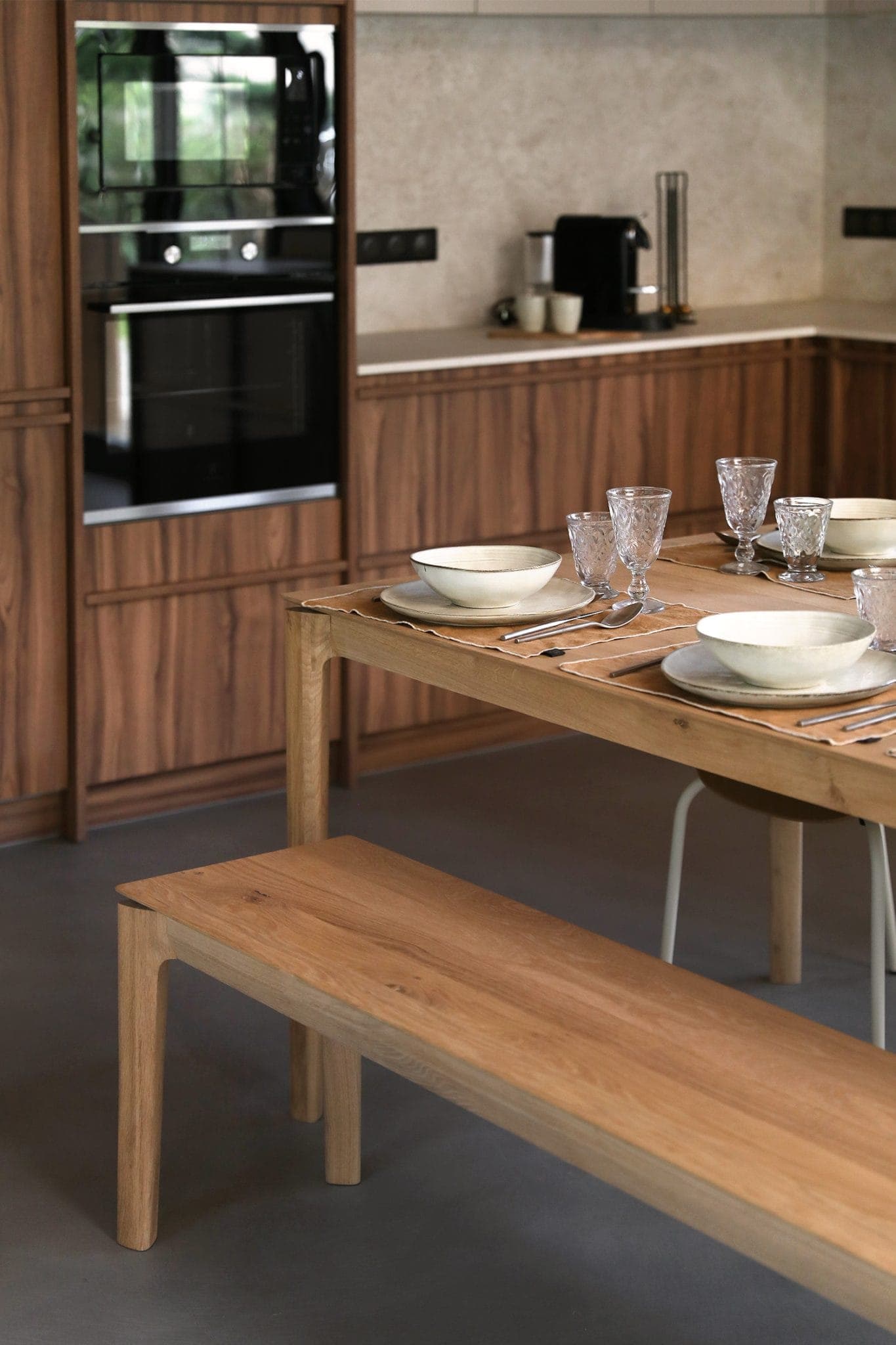 A modern kitchen with wooden cabinets and a matching dining table. The table is set with white bowls and crystal glasses on woven placemats. A bench is to one side, and a black coffee maker and oven are in the background.