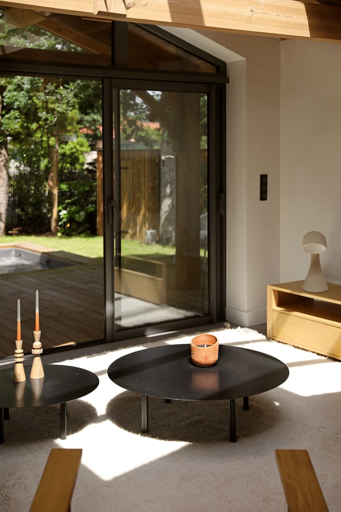 A warm living room with two round black tables, one with a candle. Wooden chairs and a small wooden shelf with a lamp are nearby. Large glass doors open onto a verdant garden, filling the room with natural light.