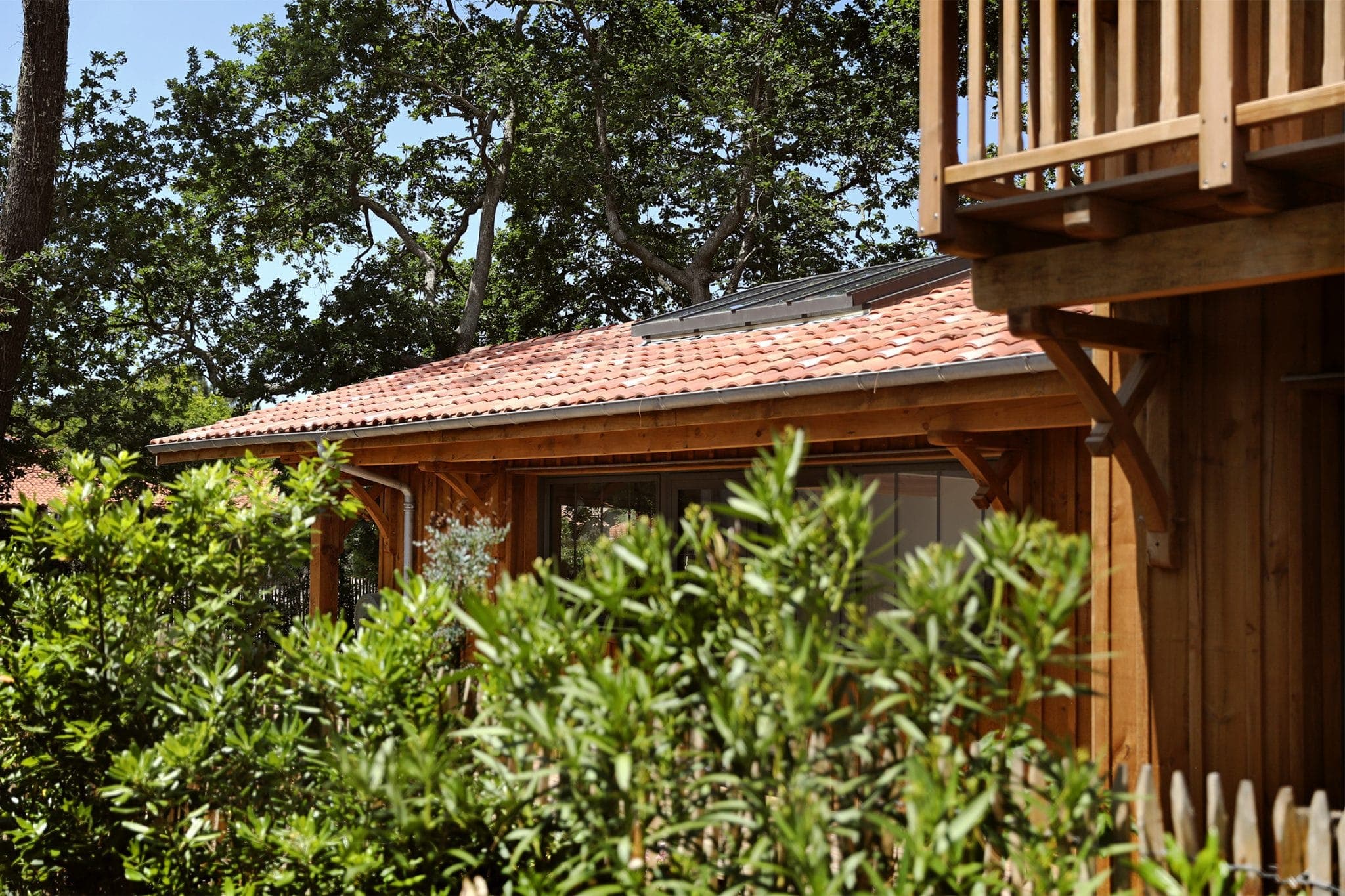 A wooden house surrounded by lush vegetation is shown. The roof is covered with red tiles and there is a wooden balcony on the right. Sunlight highlights the natural setting, with tall trees in the background under a clear blue sky.