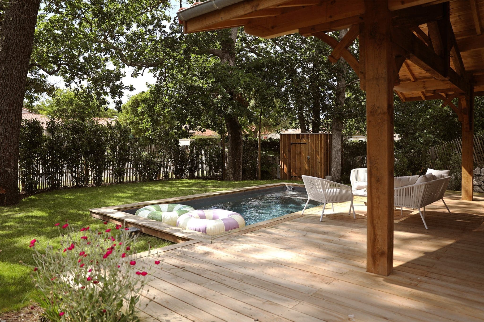 A wooden terrace with chairs overlooks a small rectangular pool with colorful inflatable rings. Surrounded by lush vegetation and tall trees, the area offers a serene, welcoming outdoor space.