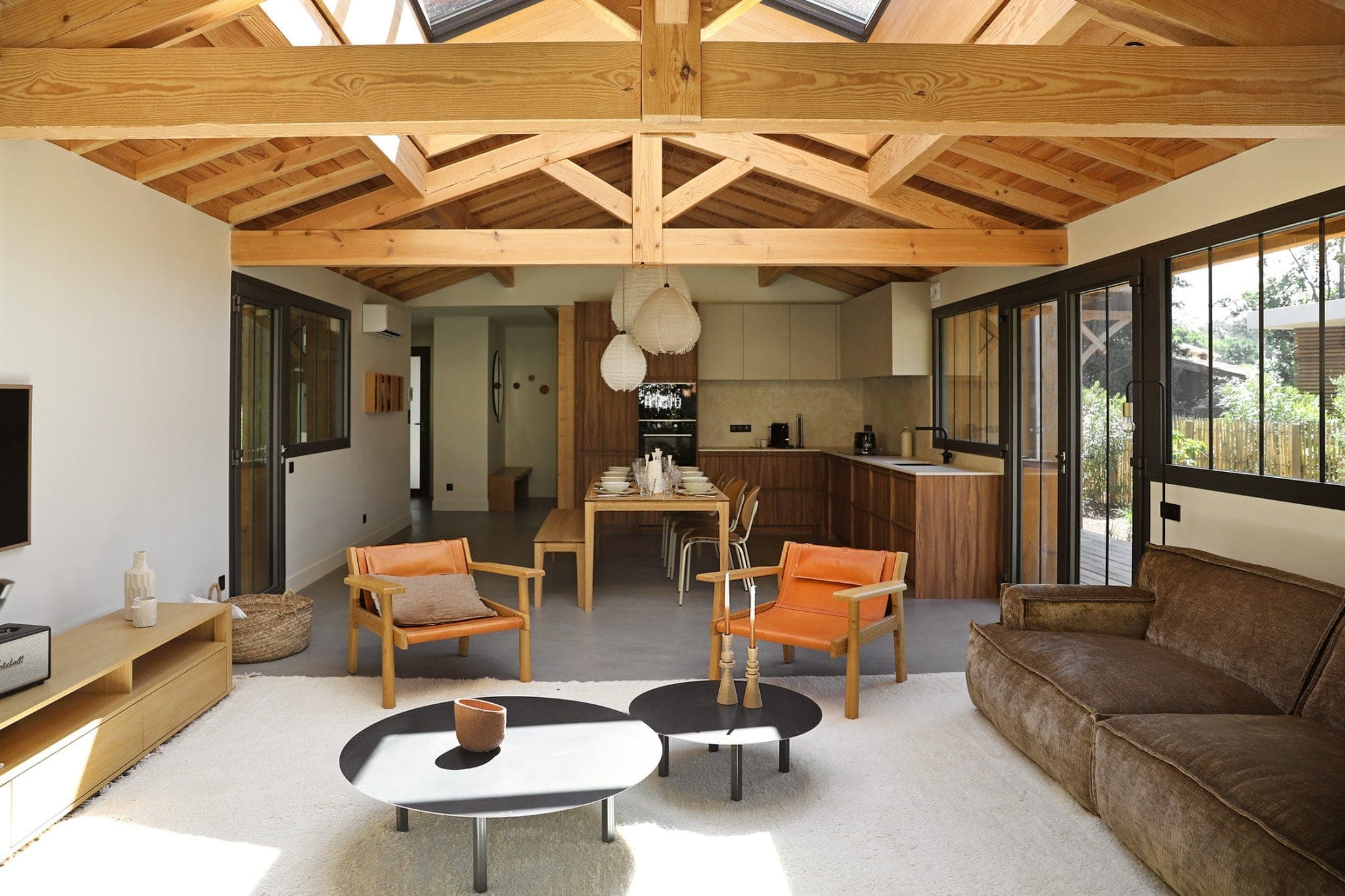 Un salon moderne avec des poutres en bois et des lucarnes. Il comprend un canapé marron, deux chaises beiges et une paire de tables basses noires sur un tapis blanc. À l'arrière-plan, on aperçoit une cuisine ouverte avec des armoires en bois et une table à manger.