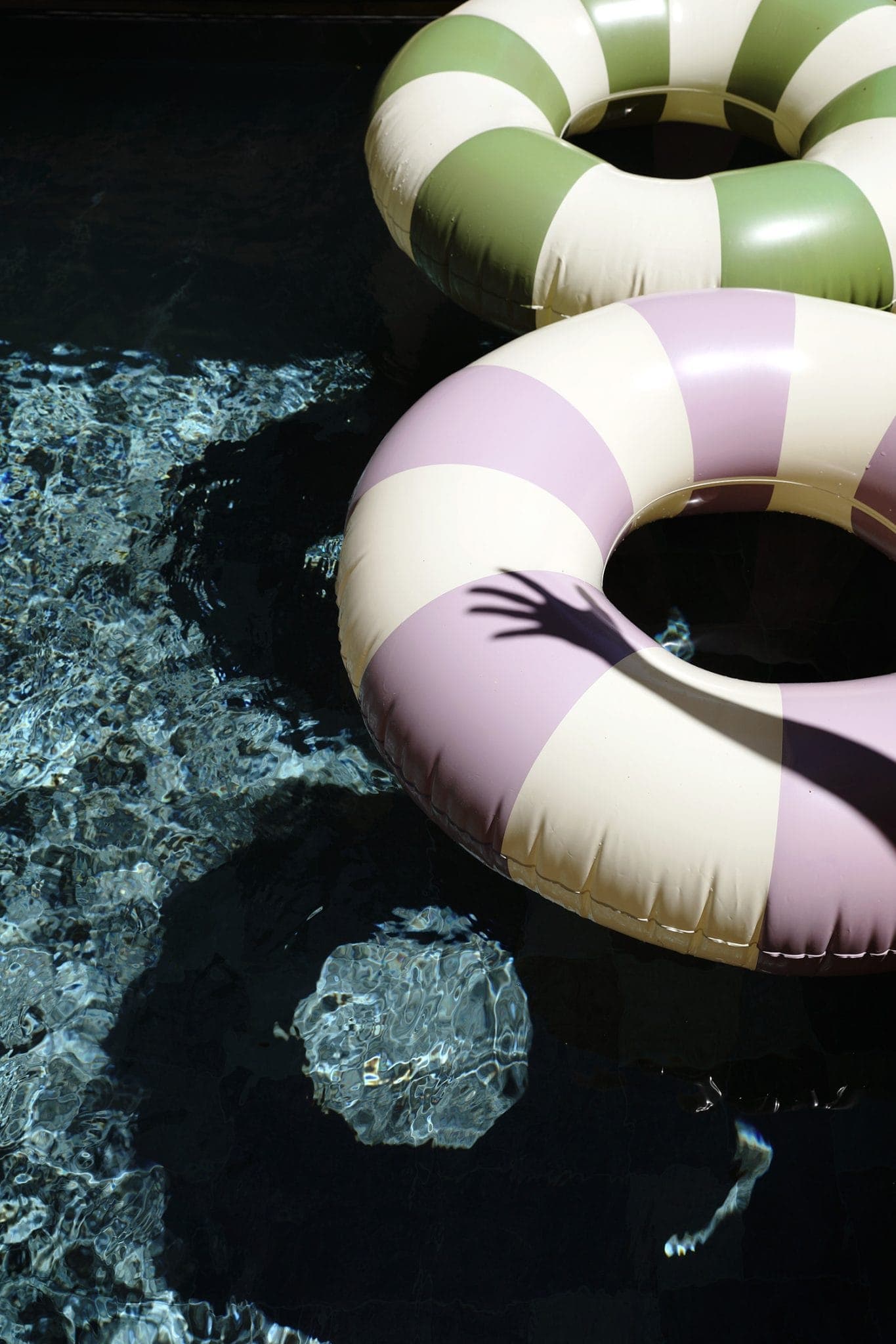 Two inflatable buoys, one with pink and white stripes, the other with green and white stripes, float on the clear water. The shadow of a hand reaches out towards them, creating an interesting pattern on the pool surface.