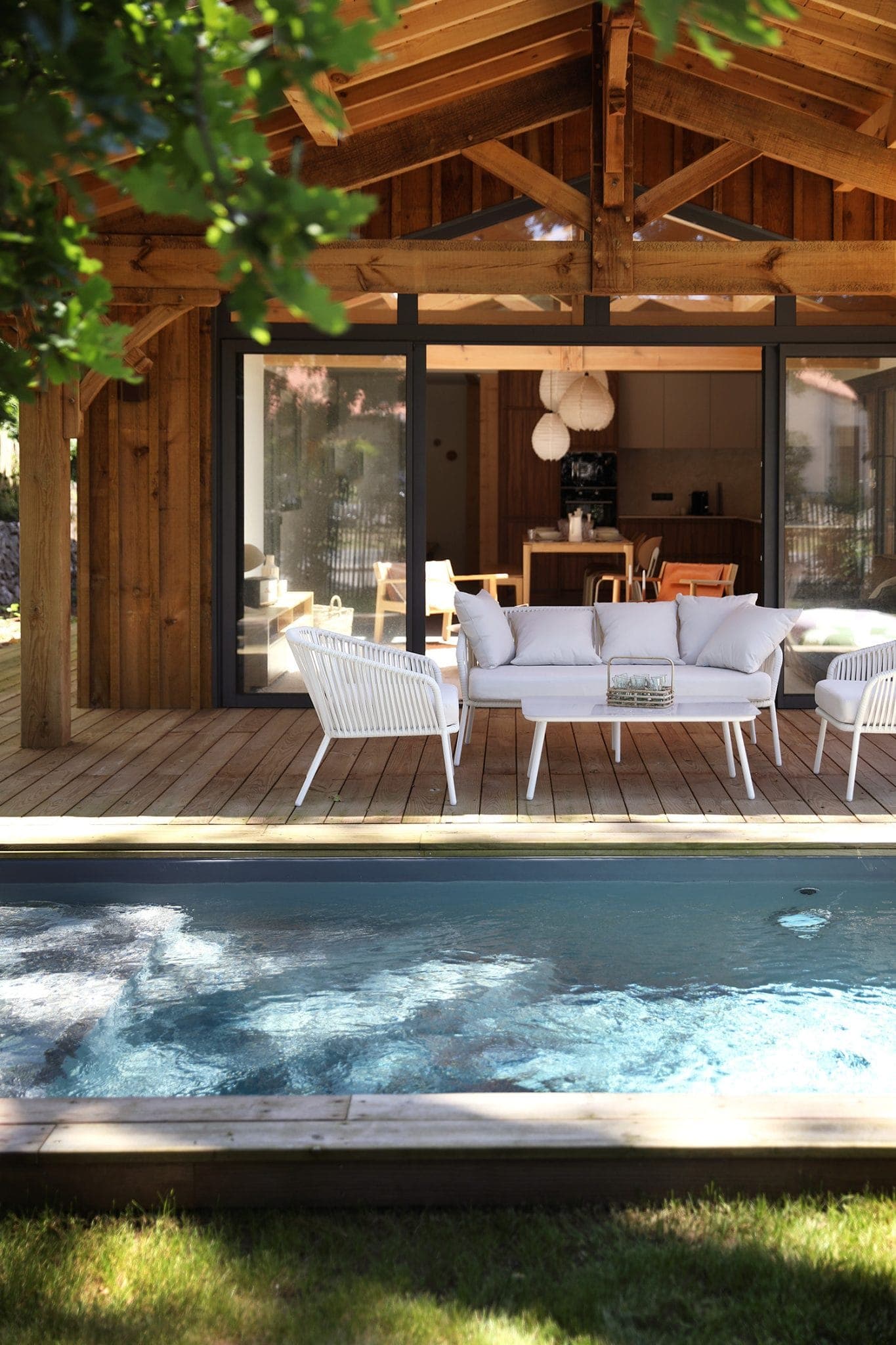 A comfortable wooden patio with white outdoor furniture stands in front of a sliding glass door, leading to a modern interior. A serene rectangular pool is in the foreground, surrounded by greenery and a wooden deck.