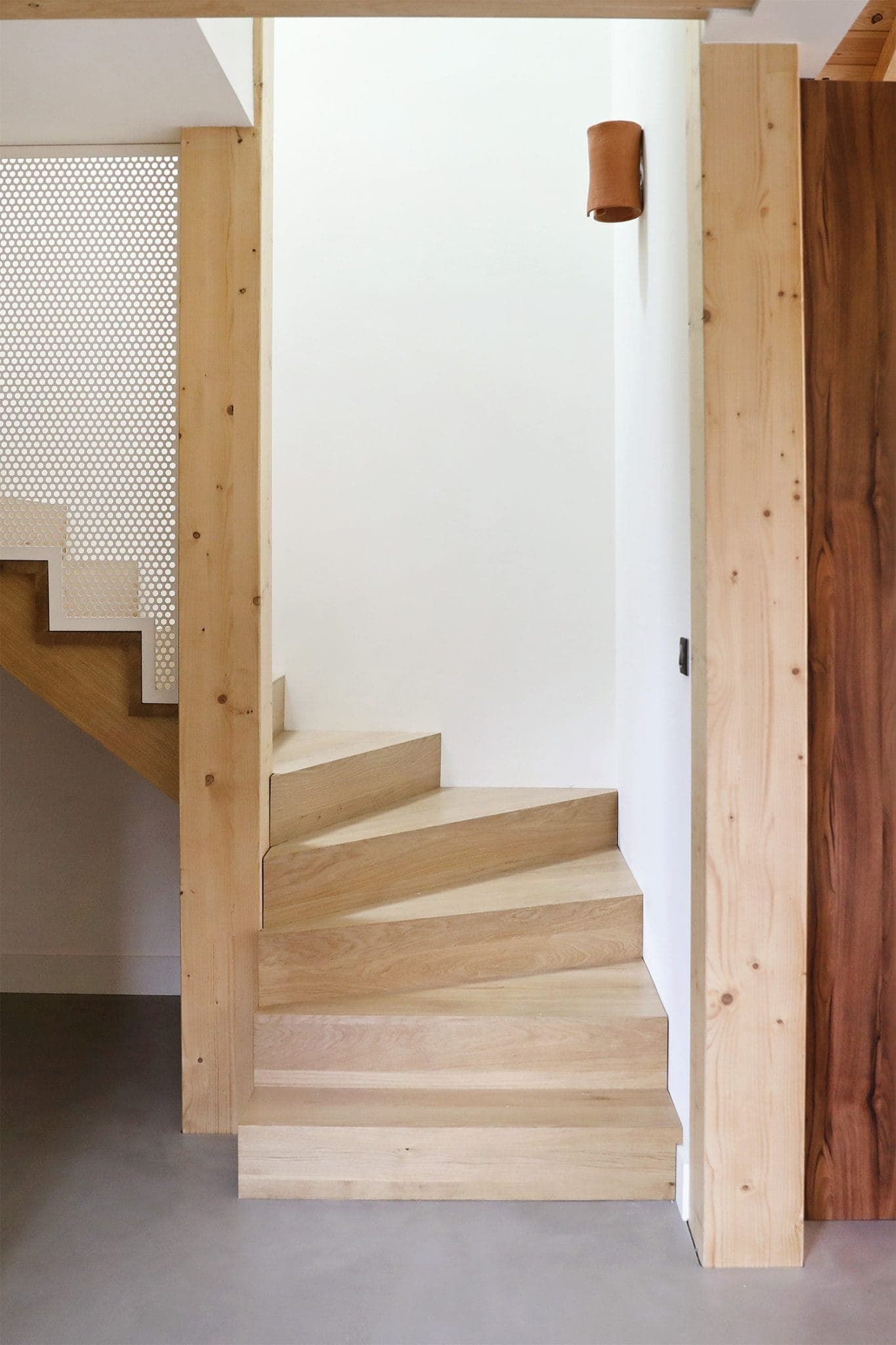 A wooden staircase with light-colored treads curves to the right, framed by wooden beams. A small wall light is mounted above, illuminating the space. White walls and a perforated panel add a modern touch to the minimalist design.