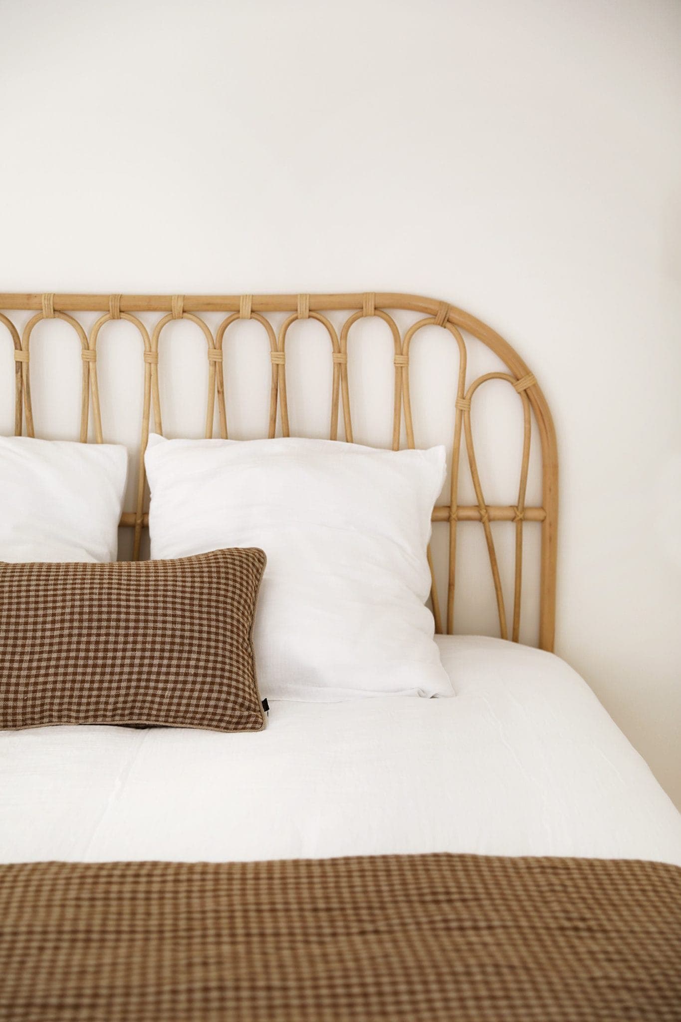 A comfortable room with a bed and rattan headboard. The bed is carefully crafted with white pillows and white sheets. A brown plaid pillow and matching blanket are placed on the bed, adding a warm touch.