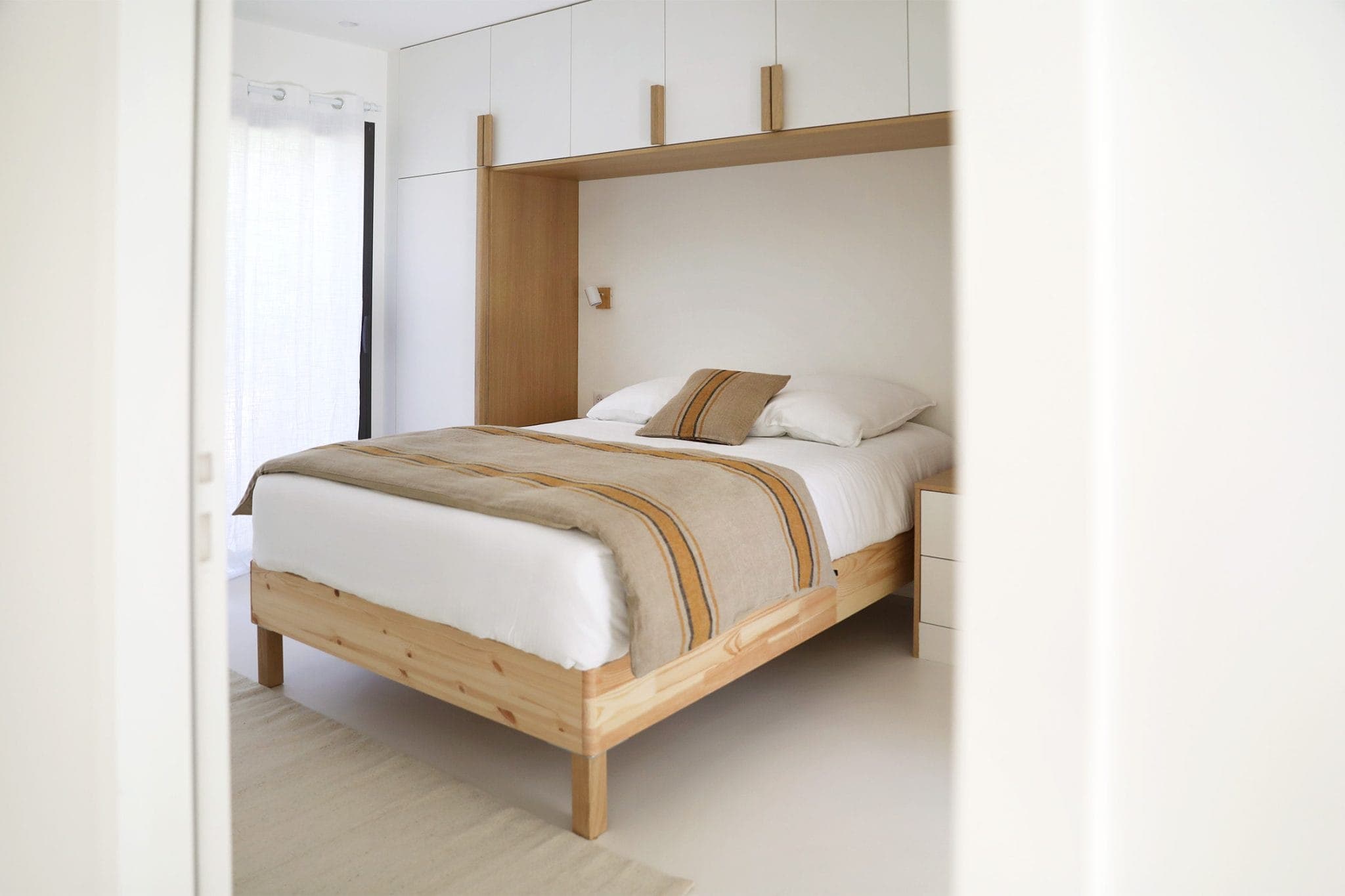 Minimalist bedroom with wooden bed, white and beige sheets, surrounded by white furniture. Natural light streams through sheer curtains, enhancing the room's clean, modern aesthetic.