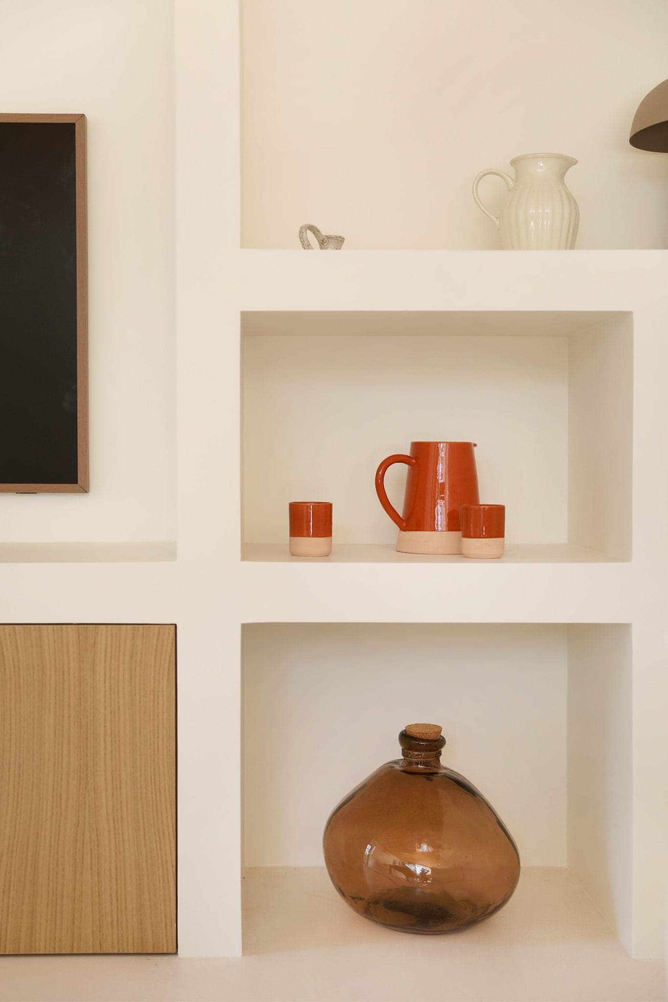 White shelf featuring a large brown glass jug on the bottom shelf and an orange pitcher with matching cups on the middle shelf. A white vase and a small figurine sit on the top shelf. Next to it is a wooden cupboard.