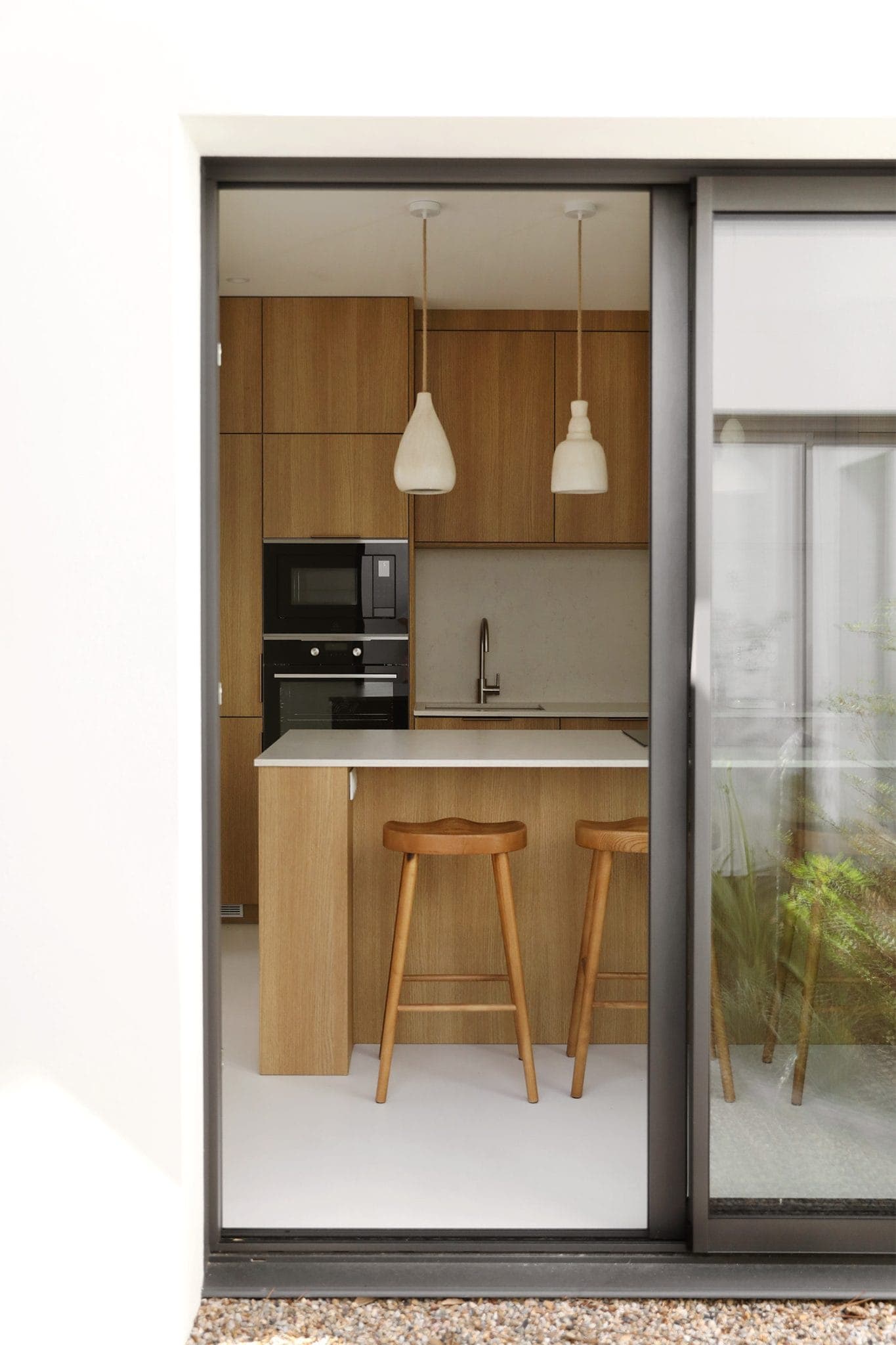 A modern kitchen with wooden cabinets, a black microwave and a white countertop. Two wooden stools are placed on the countertop. Two pendant lights hang above. The space is visible through a large sliding glass door.