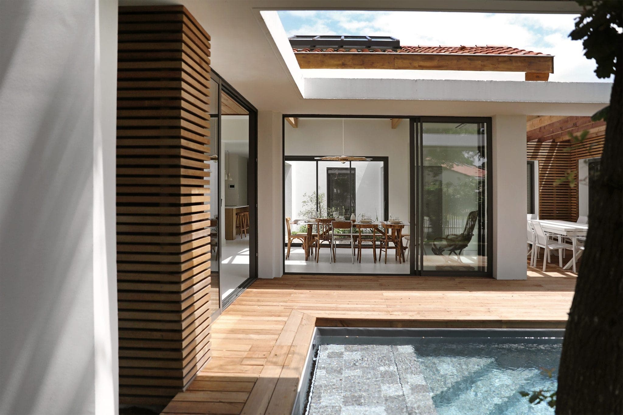 A modern house with a wooden terrace and a small swimming pool. Large glass doors reveal an indoor dining room with wooden table and chairs. The open design allows natural light to flood the space, highlighting the clean lines and contemporary architecture.