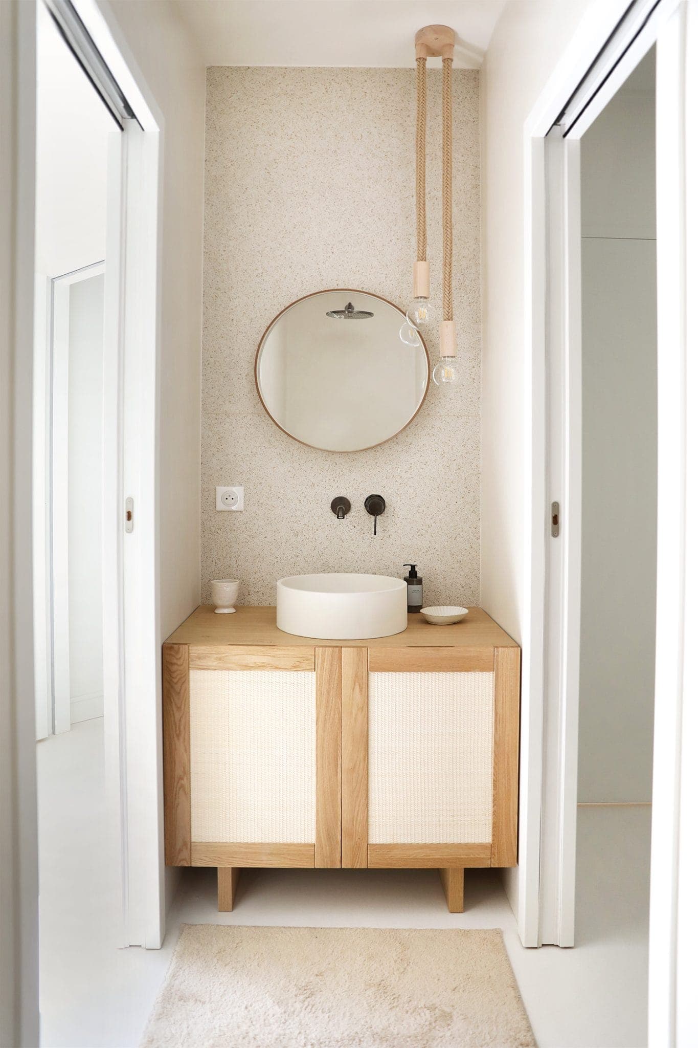 Minimalist bathroom with light wood vanity, round mirror and basin. Suspended light bulbs add a modern touch. Neutral tones and textured walls create a calm atmosphere. Doors are visible from either side.