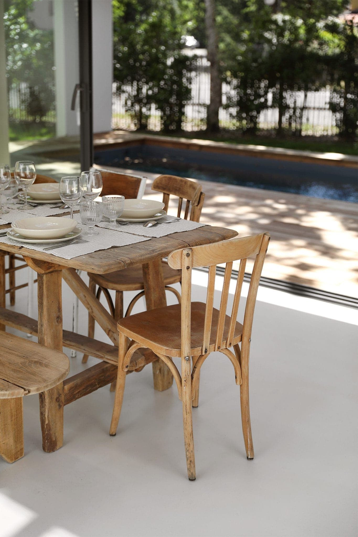 A dining table with plates, glasses and table runner is set inside near a large glass window. Outside, there's a view of a swimming pool and the greenery beyond. Wooden chairs surround the rustic table.