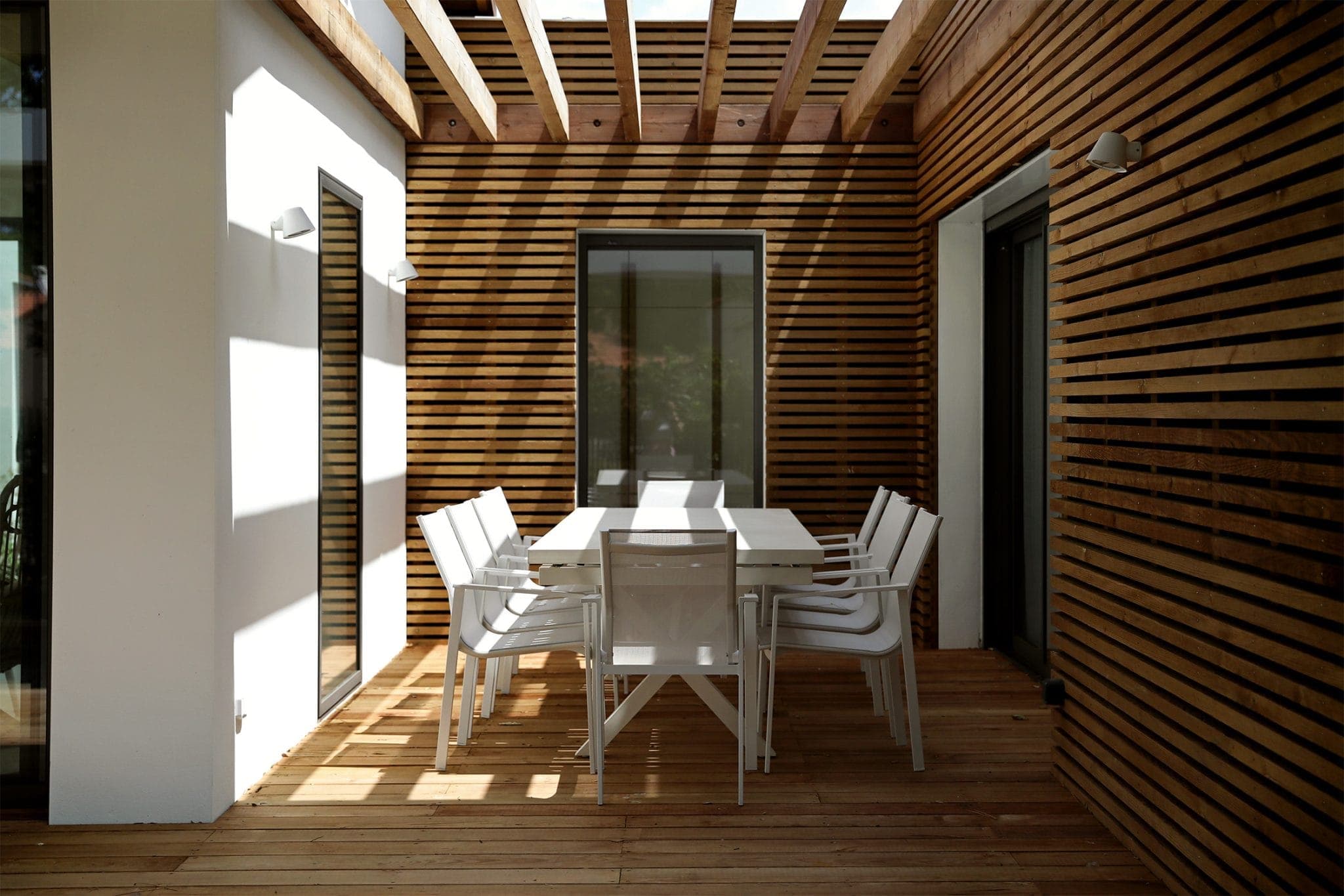 A minimalist outdoor dining area with a long white table and eight white chairs. The space features wooden slatted walls, a wooden floor and a pergola allowing sunlight to cast shadows. Two large glass doors are visible.