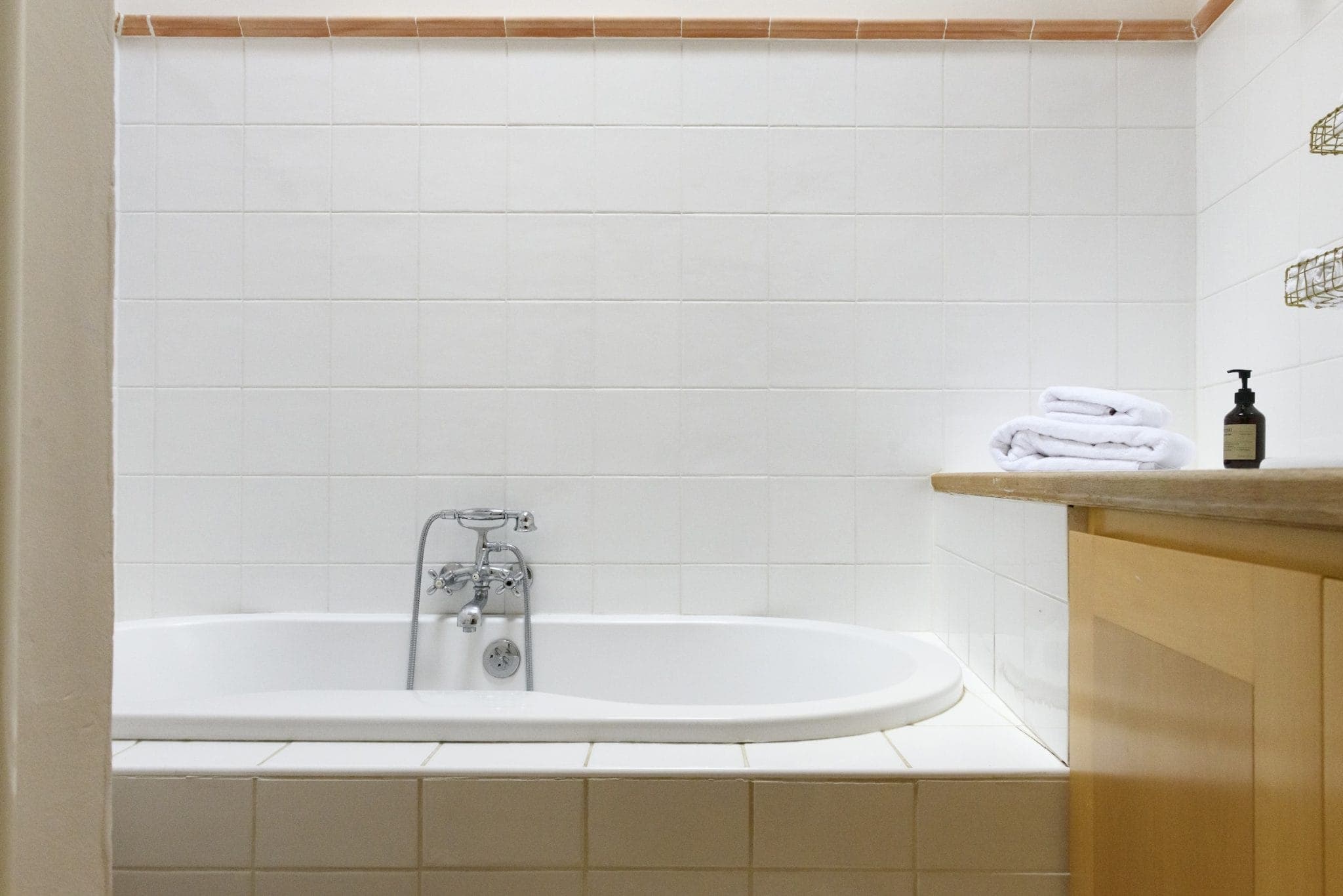 A minimalist bathroom features a white bathtub with vintage-style accessories against a tiled wall. On a nearby wooden countertop, folded white towels and a soap dispenser rest neatly. The atmosphere is clean and serene.