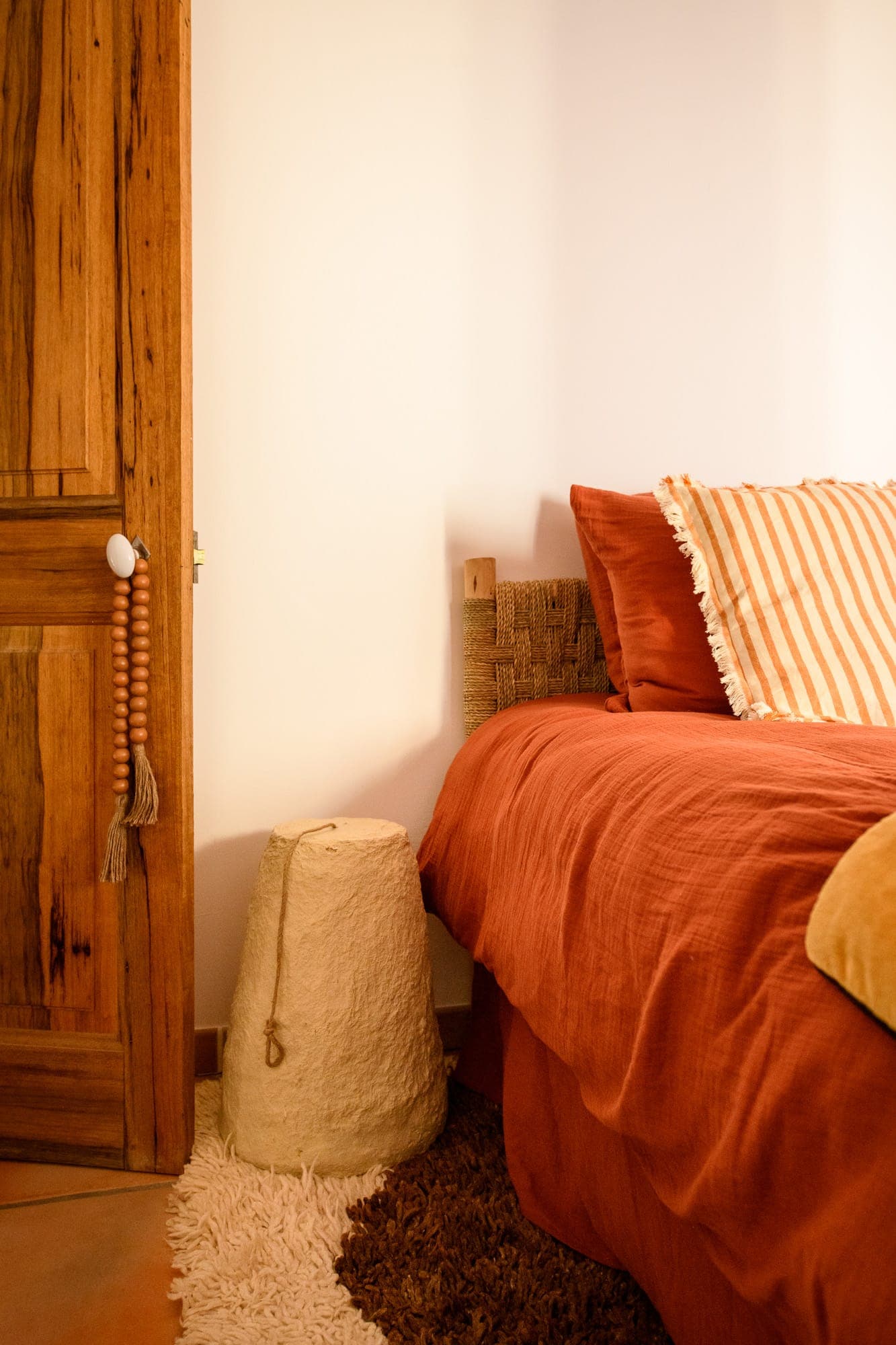 Coin chambre cosy à l'ambiance rustique. Il comprend un couvre-lit orange, des oreillers rayés, une tête de lit en bois et une porte en bois. Une lampe texturée repose sur un tapis moelleux à côté du lit, complétant les tons terreux.