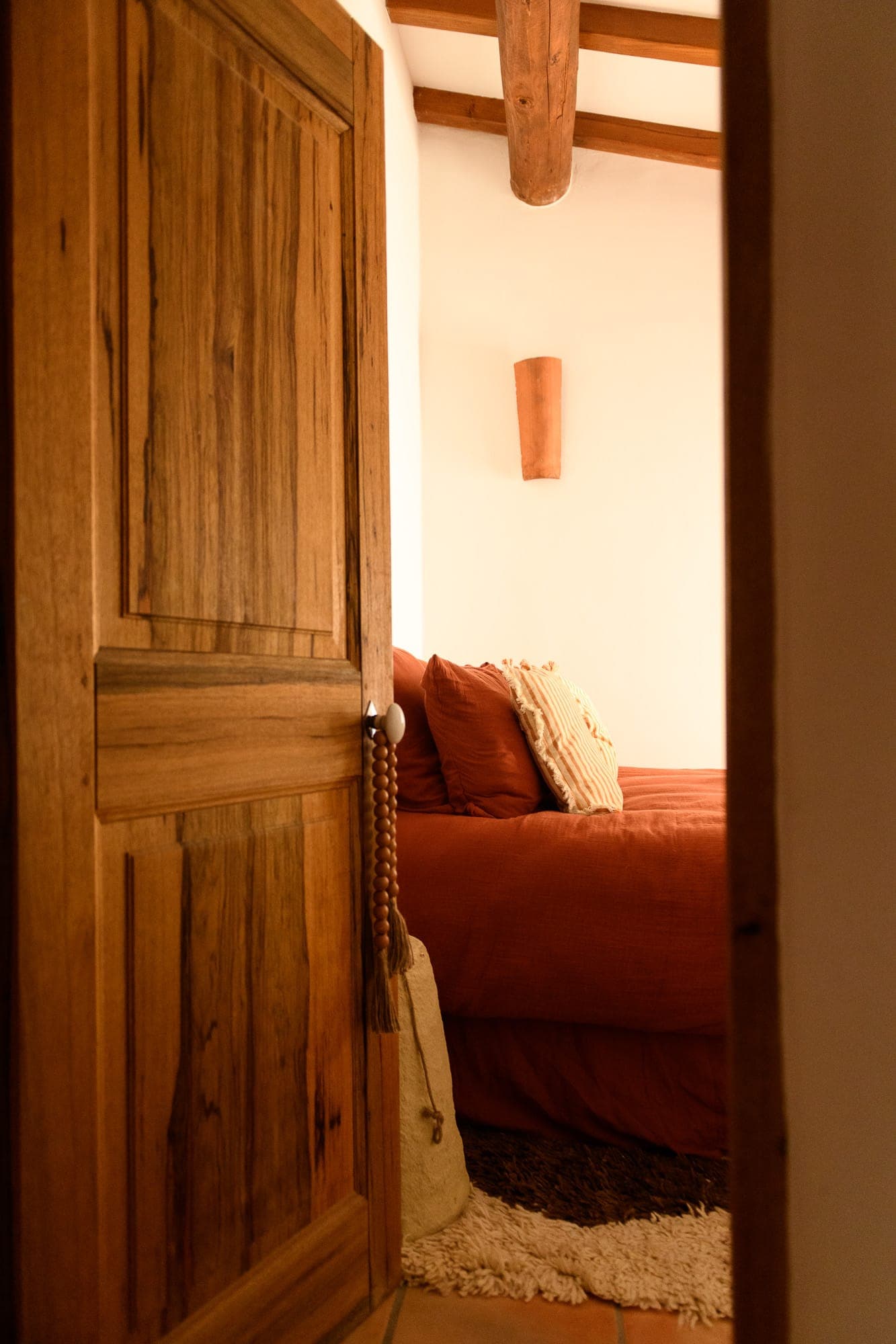 Une chambre confortable vue à travers une porte en bois partiellement ouverte. La chambre dispose d'un lit avec une literie couleur rouille, d'un oreiller rayé, d'un tapis moelleux et d'un éclairage chaleureux, créant une atmosphère accueillante et rustique.