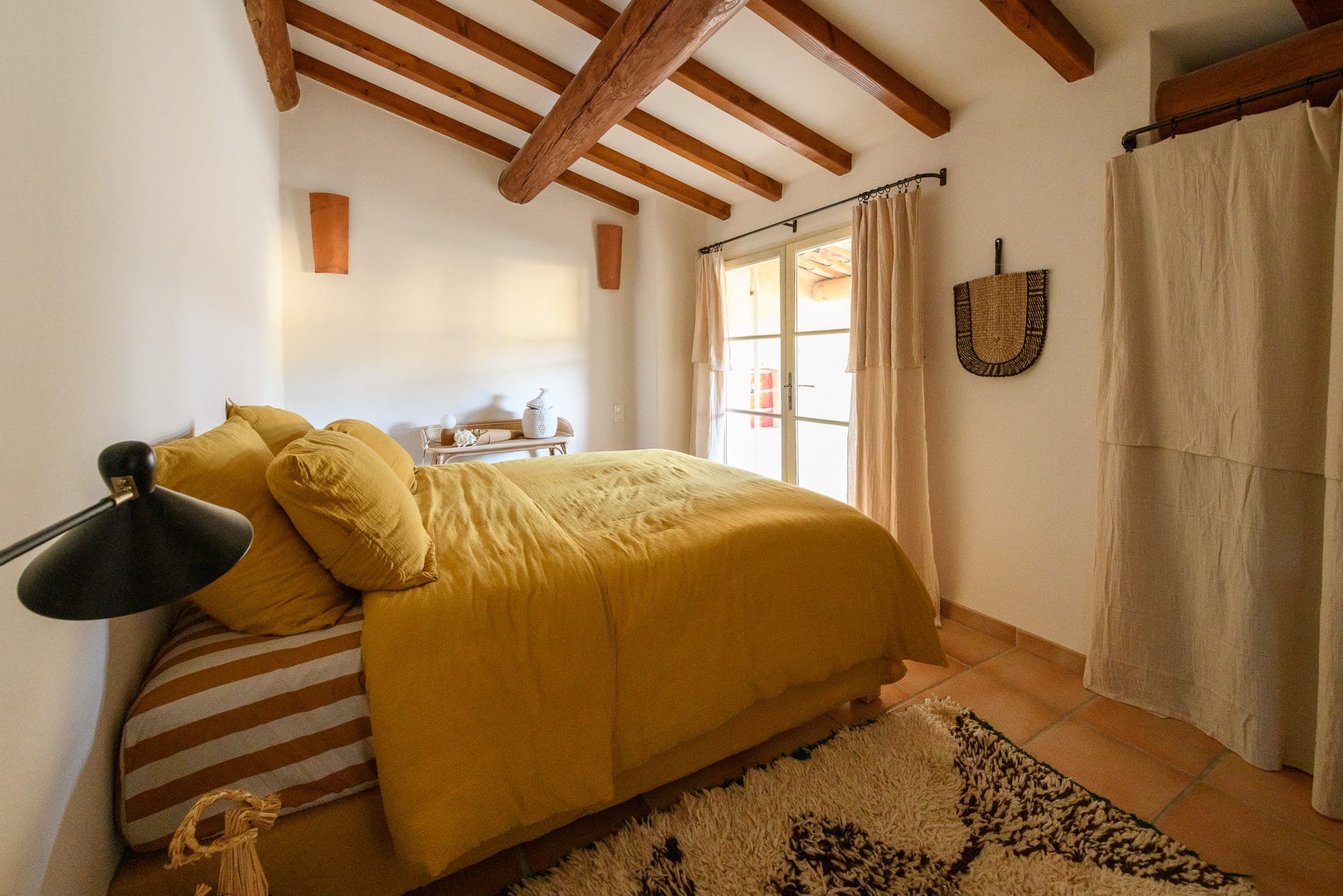 Une chambre confortable avec un plafond à poutres en bois et un lit habillé de draps jaunes. La lumière naturelle pénètre à travers une porte vitrée encadrée de rideaux légers. Une petite table d'appoint accueille des objets décoratifs et une tenture murale tissée ajoute de la texture.