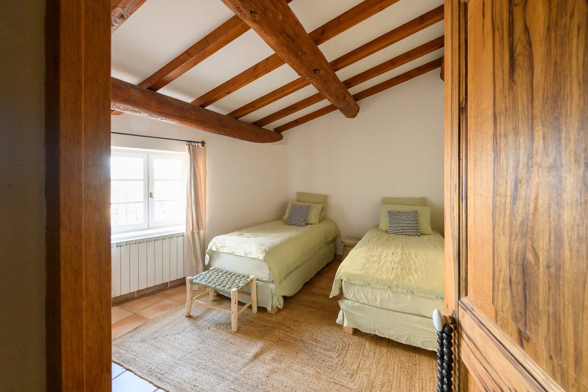 Une chambre confortable avec deux lits simples avec une literie vert clair, des poutres en bois au plafond, une fenêtre avec des rideaux transparents, une porte en bois et un tapis tissé au sol. Un petit banc est placé au pied d'un lit.