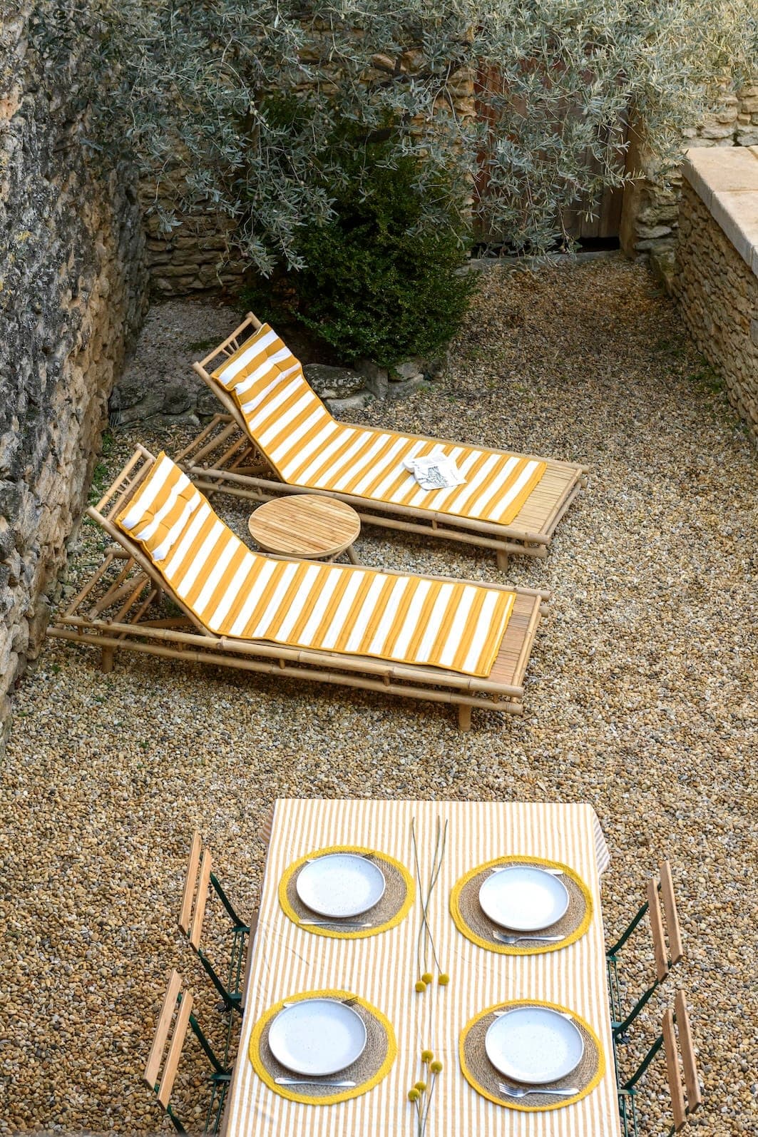 Un espace extérieur chaleureux avec deux chaises longues rayées jaune et blanc à côté d'un mur en pierre et d'une petite table ronde. Au premier plan, une table à manger est dressée avec quatre assiettes et des sets de table, en harmonie avec les couleurs des chaises longues. Le tout est entouré de galets et de verdure.