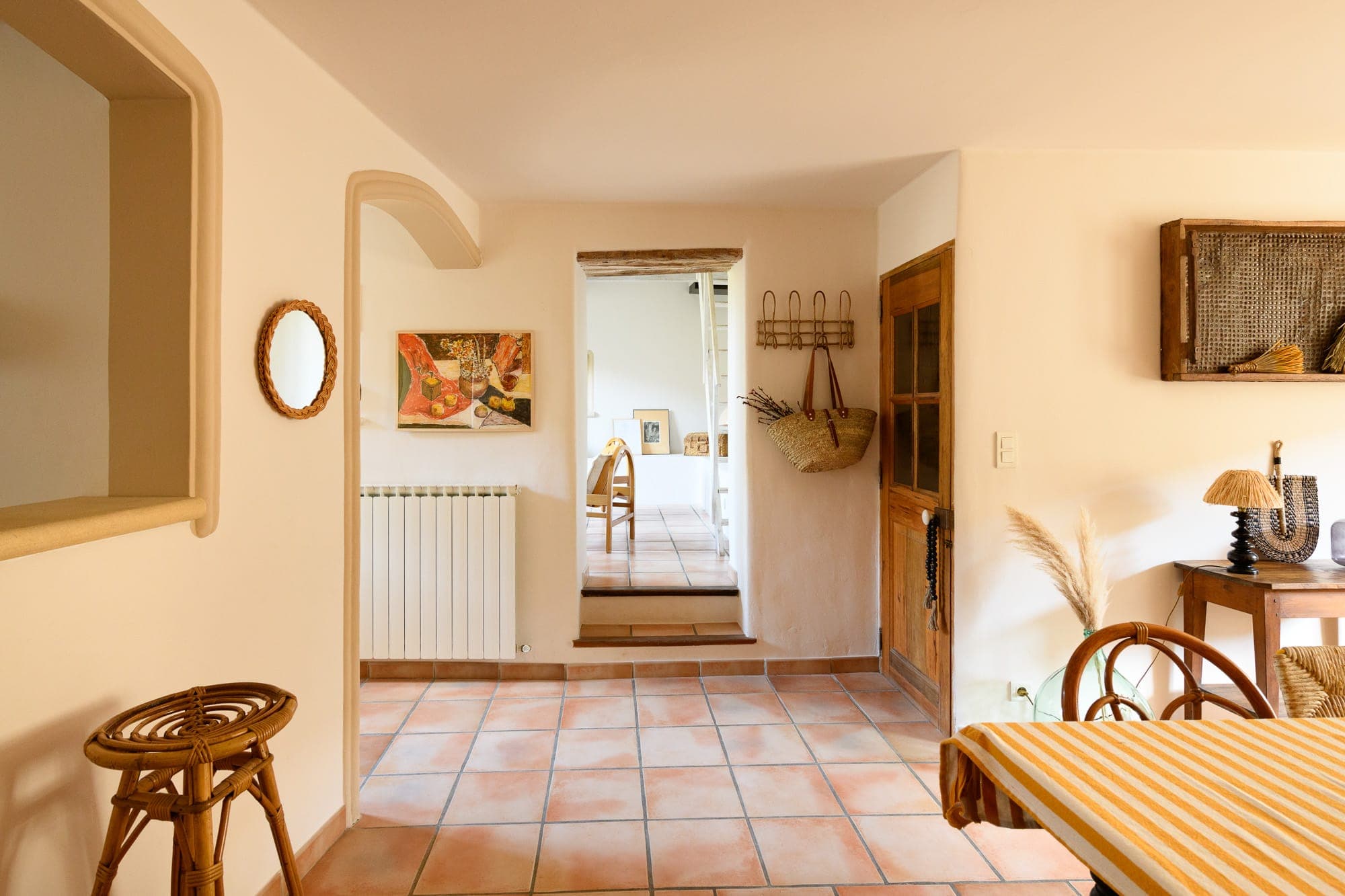 A warm and sunny dining and hallway space with terracotta tiled floors, light-colored walls, wicker furniture and decorative elements such as a wall mirror, artwork and a striped tablecloth. The space exudes a warm, rustic ambience.