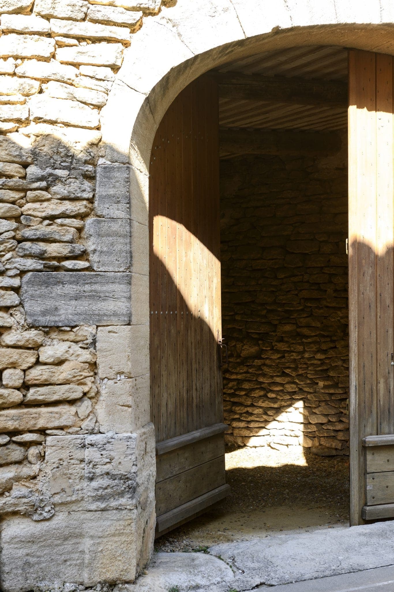 A stone arch with wooden double doors, partially open, revealing a dark interior. Sunlight casts shadows on the textured stone walls and wooden doors. The scene conveys a sense of mystery and history.