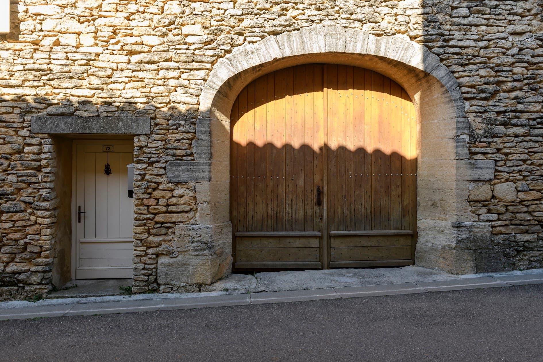 Une grande porte en bois cintrée est encastrée dans un mur de pierre rustique, à côté d'une porte blanche plus petite. La lumière du soleil projette une ombre sur la porte en bois, mettant en valeur sa surface texturée. Une rue pavée passe devant le bâtiment.