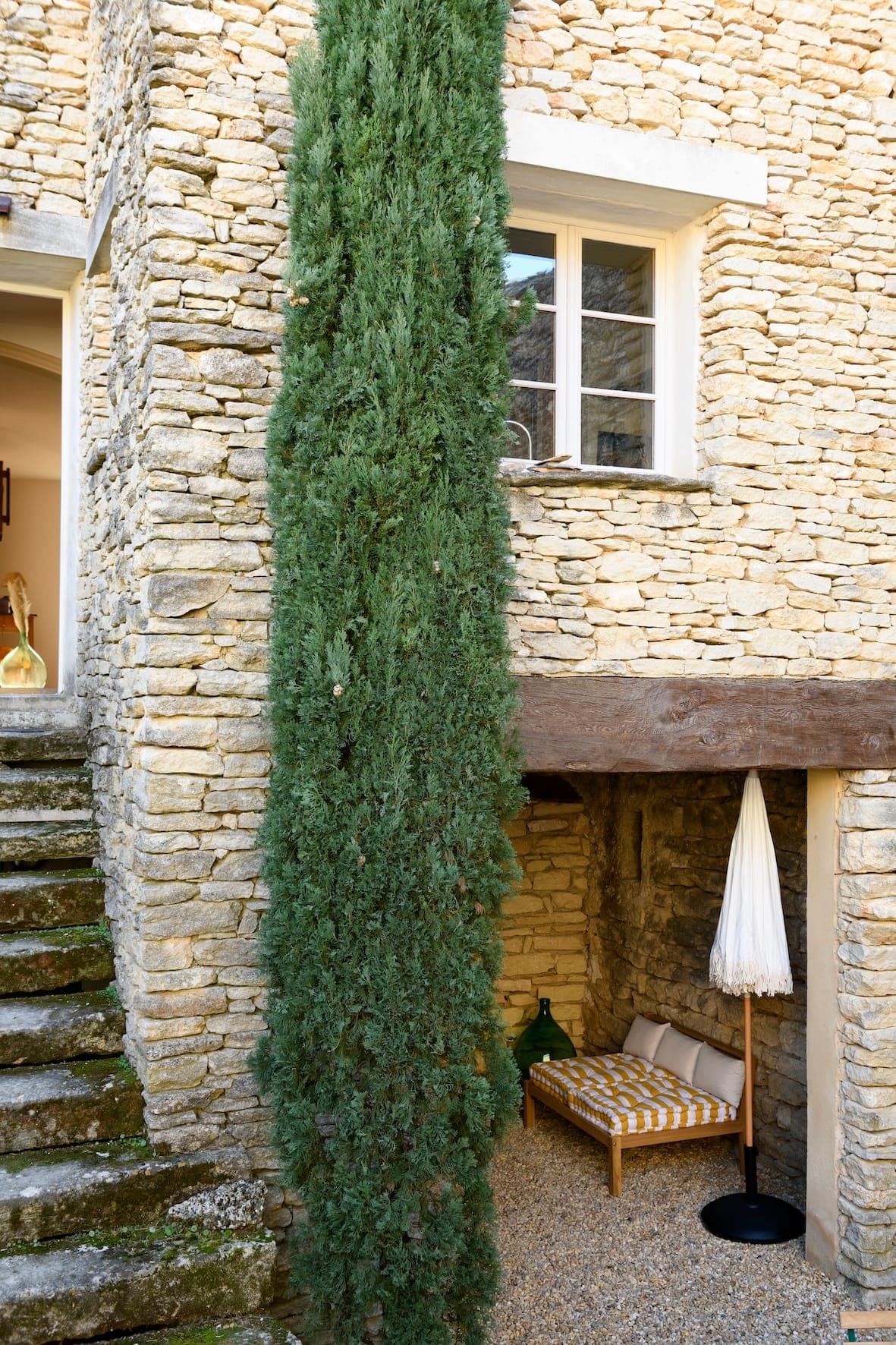 Un grand et étroit arbre à feuilles persistantes se dresse à côté d'un bâtiment en pierre. Sous une fenêtre, un petit patio est doté d'un banc rembourré avec une housse rayée et d'un parasol blanc fermé. Des marches en pierre mènent à une entrée sur la gauche.
