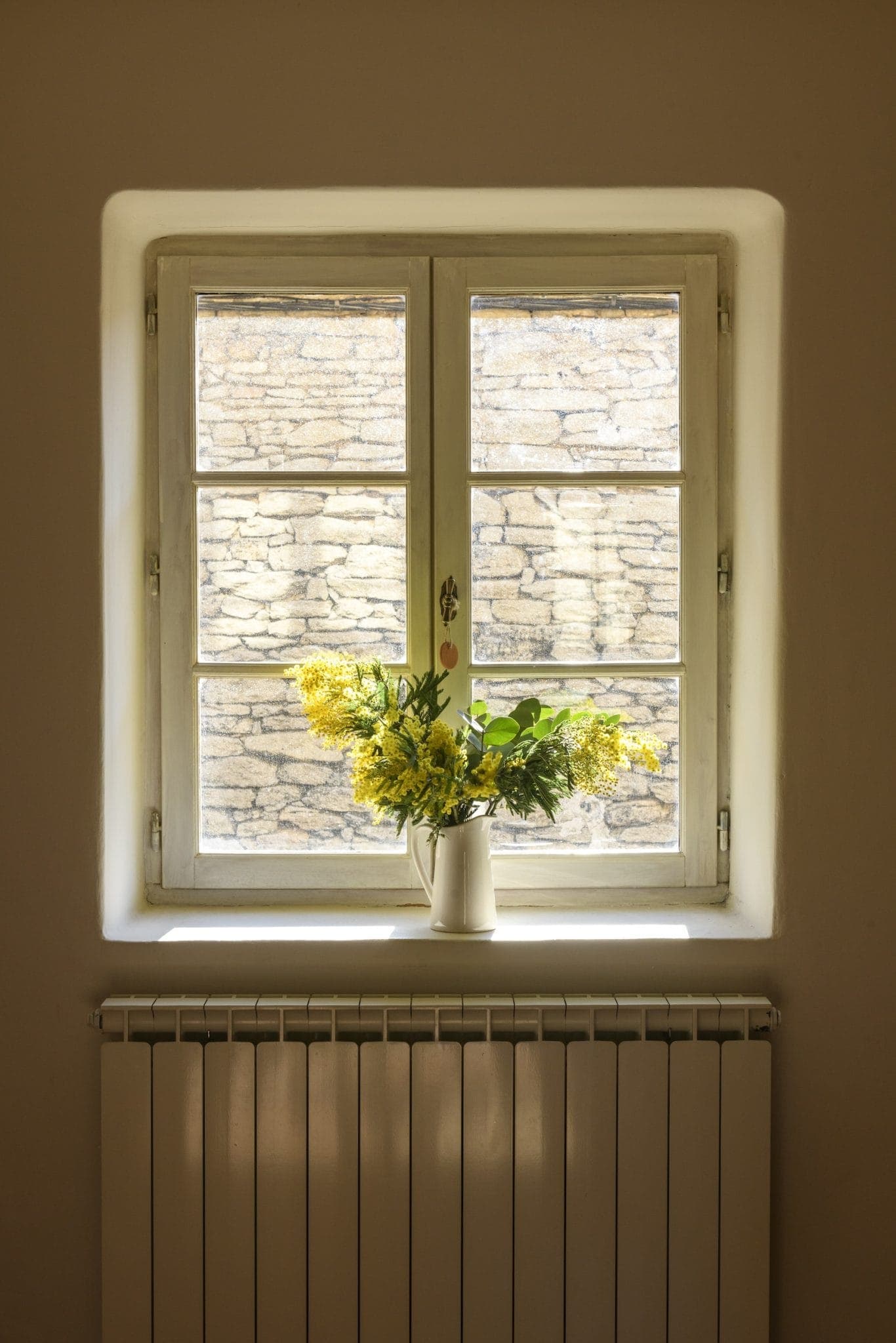 A four-paned window lets in sunlight, illuminating a white pitcher filled with yellow flowers on the windowsill. The background shows a textured brick wall, and a white radiator is visible beneath the window.