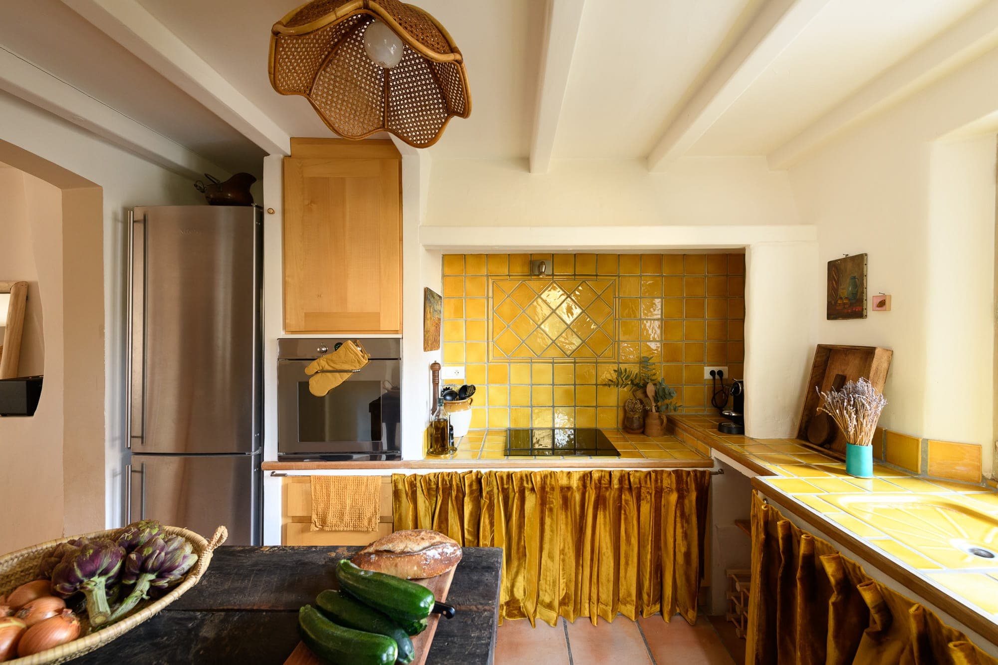 Cuisine chaleureuse de style rustique, avec dosseret et comptoirs en carrelage jaune. Un réfrigérateur en acier inoxydable se trouve à côté d'armoires en bois. L'îlot est doté de légumes frais et un luminaire en osier est suspendu au-dessus. Un panier d'artichauts est au premier plan.