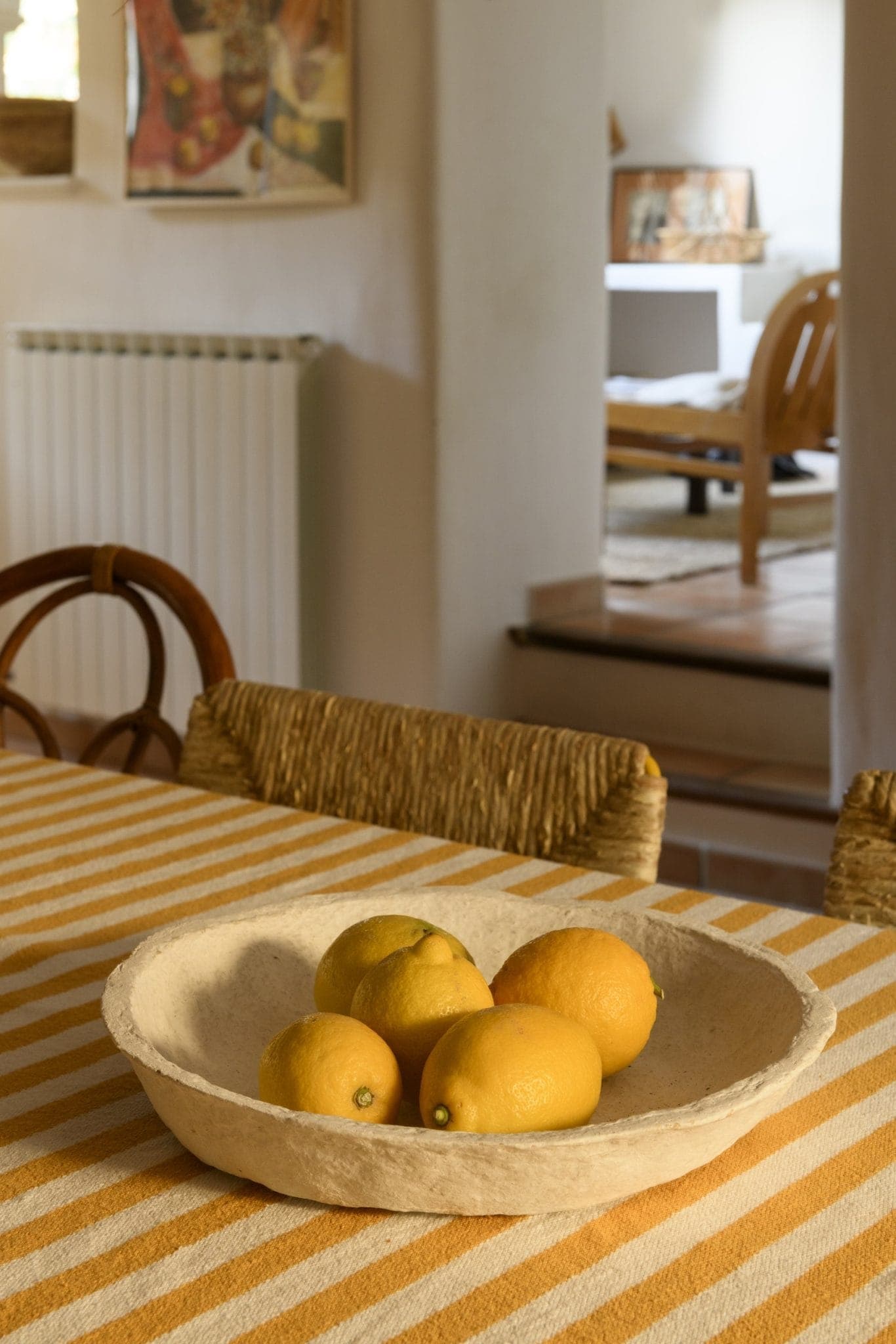 A rustic dining room features a yellow and white striped tablecloth with a ceramic bowl filled with lemons in the center. Wicker chairs surround the table, and a cozy living space is visible in the background. Warm, natural light fills the room.