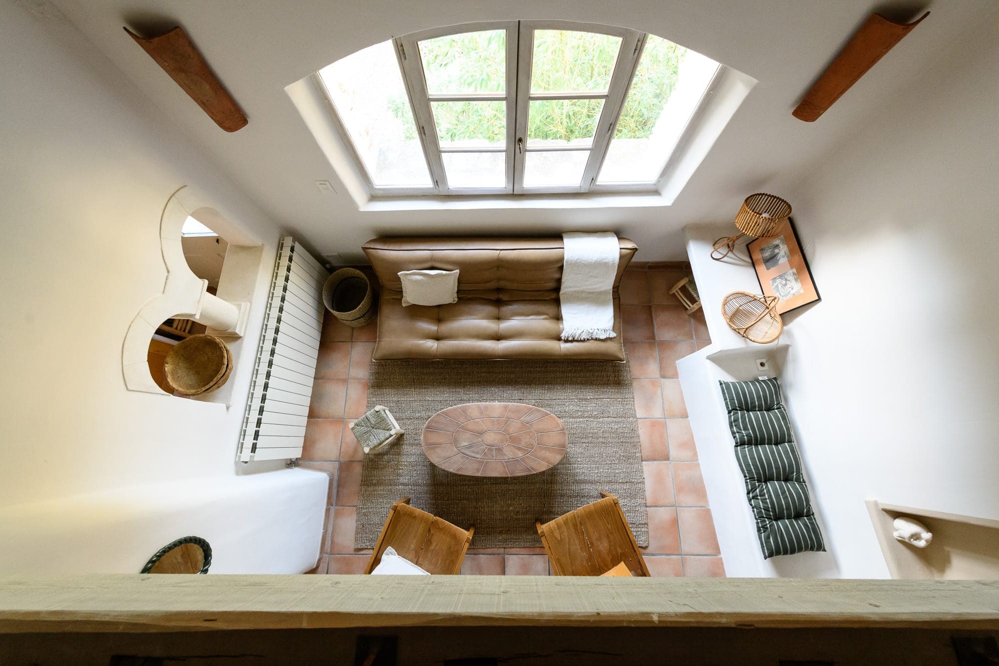 A warm living room with brown leather sofa, wooden coffee table, wicker chairs and radiator. A large arched window lets in natural light. The floor is tiled and various decorative objects are carefully arranged. View from above.