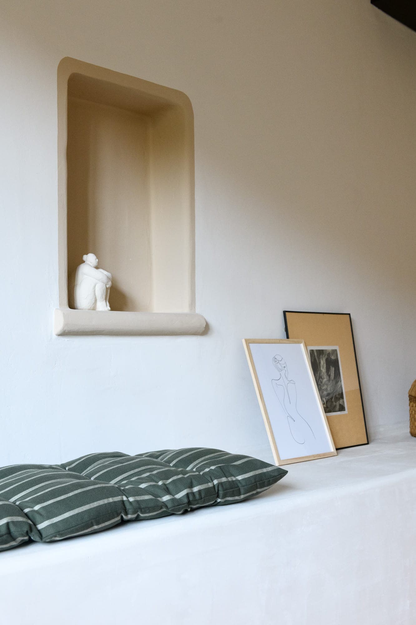 A minimalist interior with a small sculpture in a niche, green striped cushions on a white bench and framed artwork leaning against the wall. The setting is calm and the aesthetic modern.