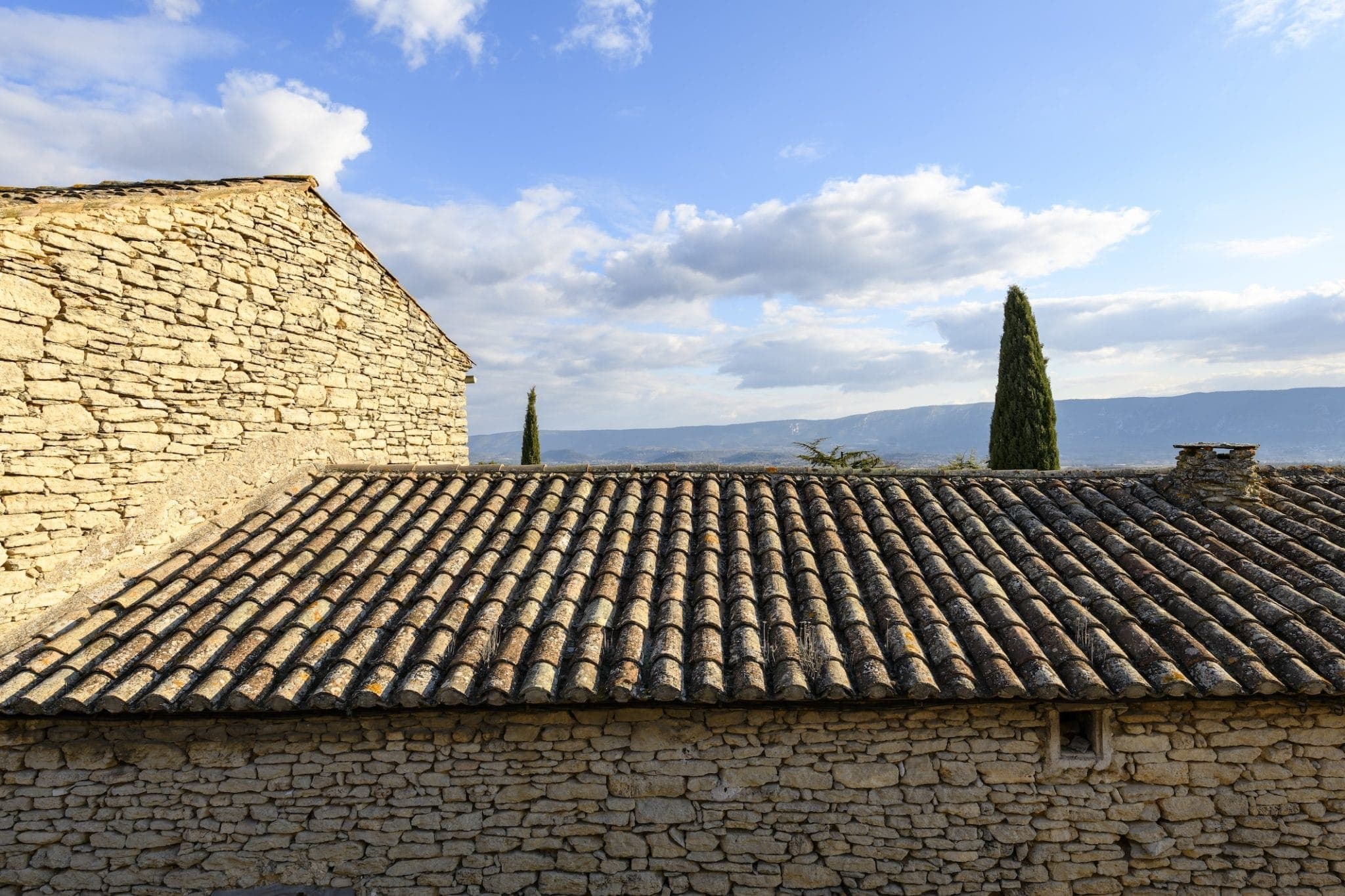 A rustic stone building with a terracotta tile roof under a blue sky dotted with clouds. Two tall cypress trees are visible in the background, with a mountainous landscape in the distance.