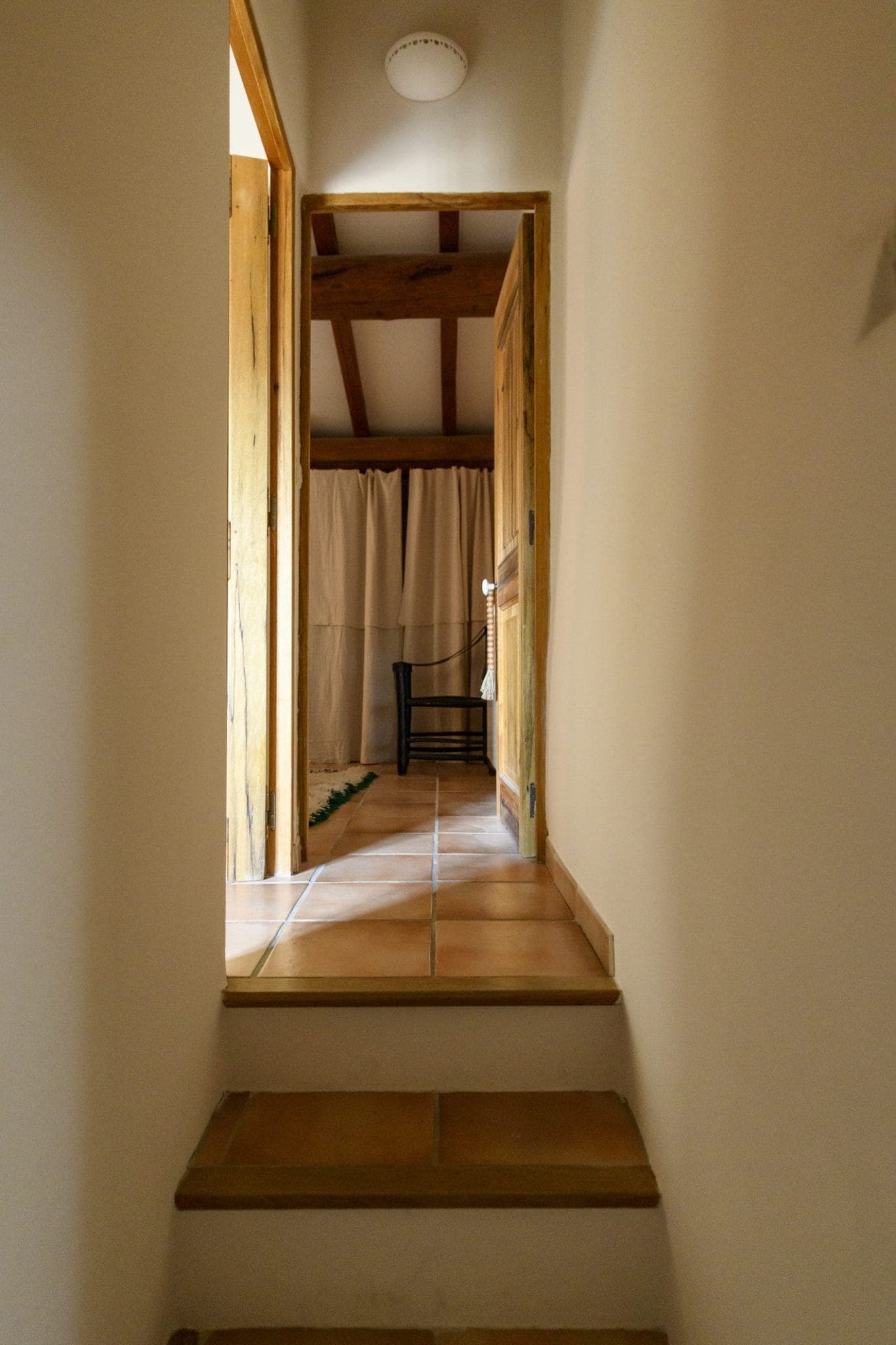 A dimly lit room with wooden beams and a tiled floor, with an open door at the top of a small staircase. A black chair with a woven seat is visible in the room, with transparent light-colored curtains behind it.
