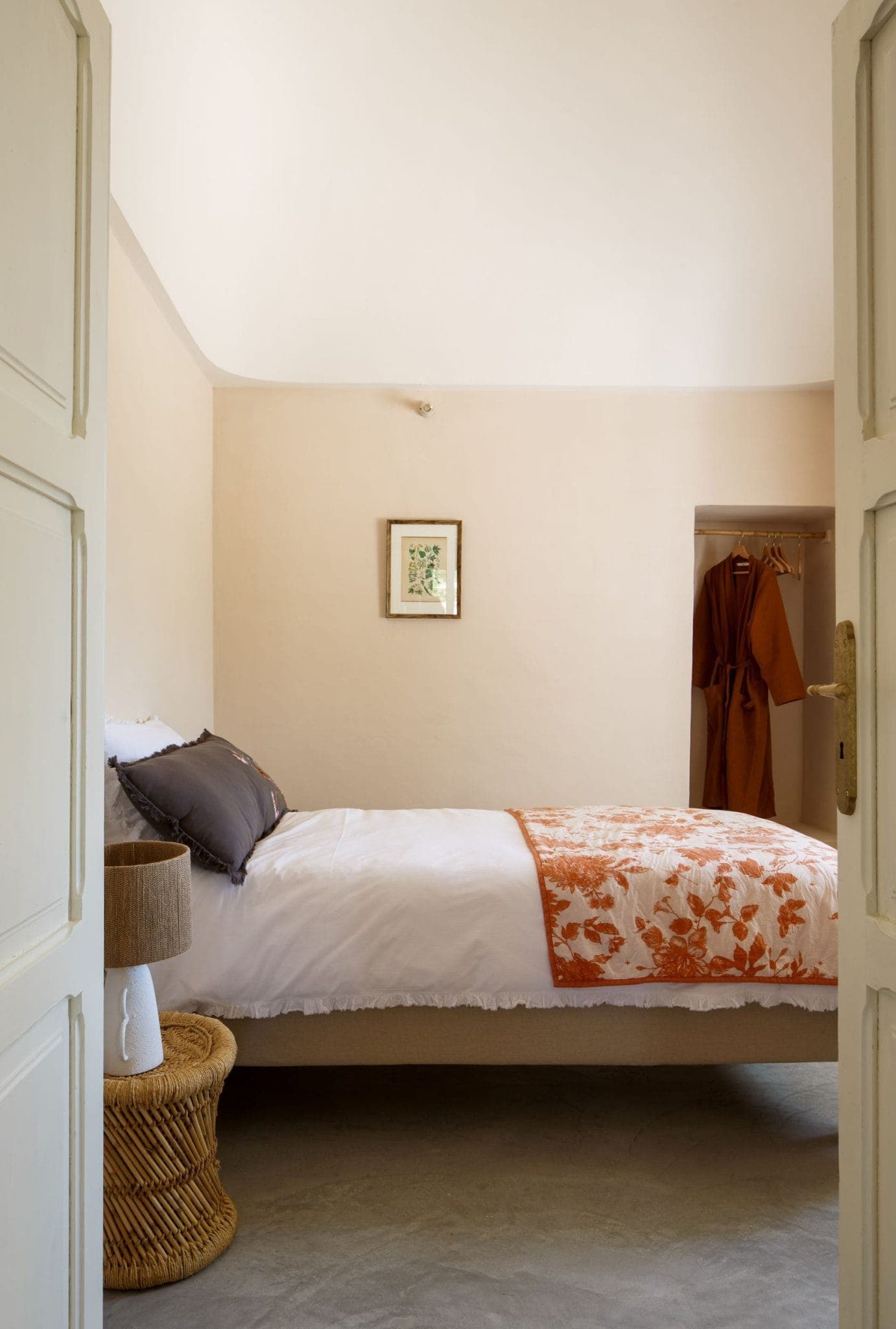 A comfortable room with a well-made bed with white and orange floral bedding. There's a small woven side table with a lamp, a closet with clothes and a framed picture on the wall. The room has an open door, neutral walls and a serene ambience.