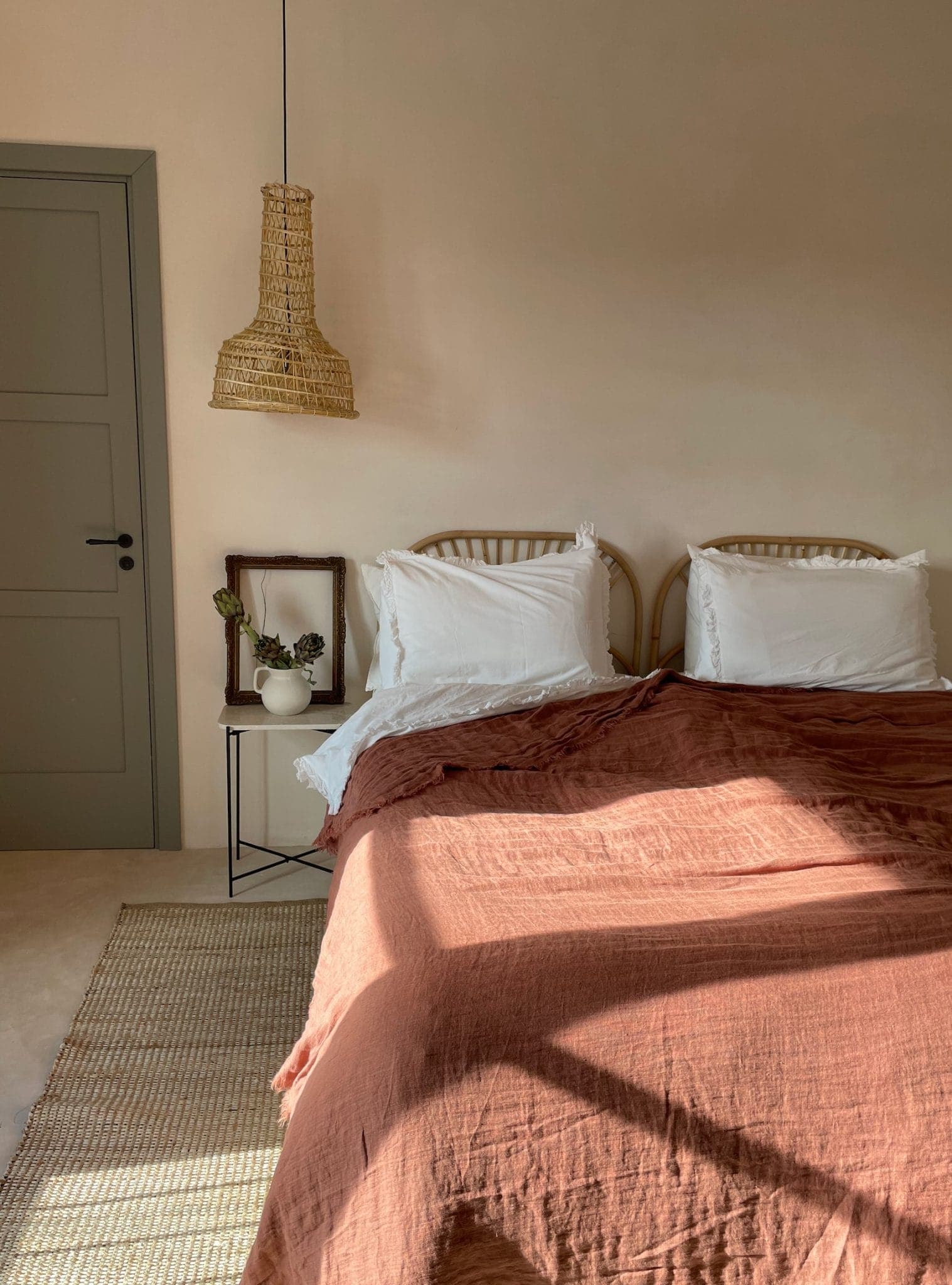 A comfortable room with a double bed with white pillows and a rust-colored blanket. A woven pendant hangs above a small side table with a vase. The room features soft lighting and a neutral color palette.