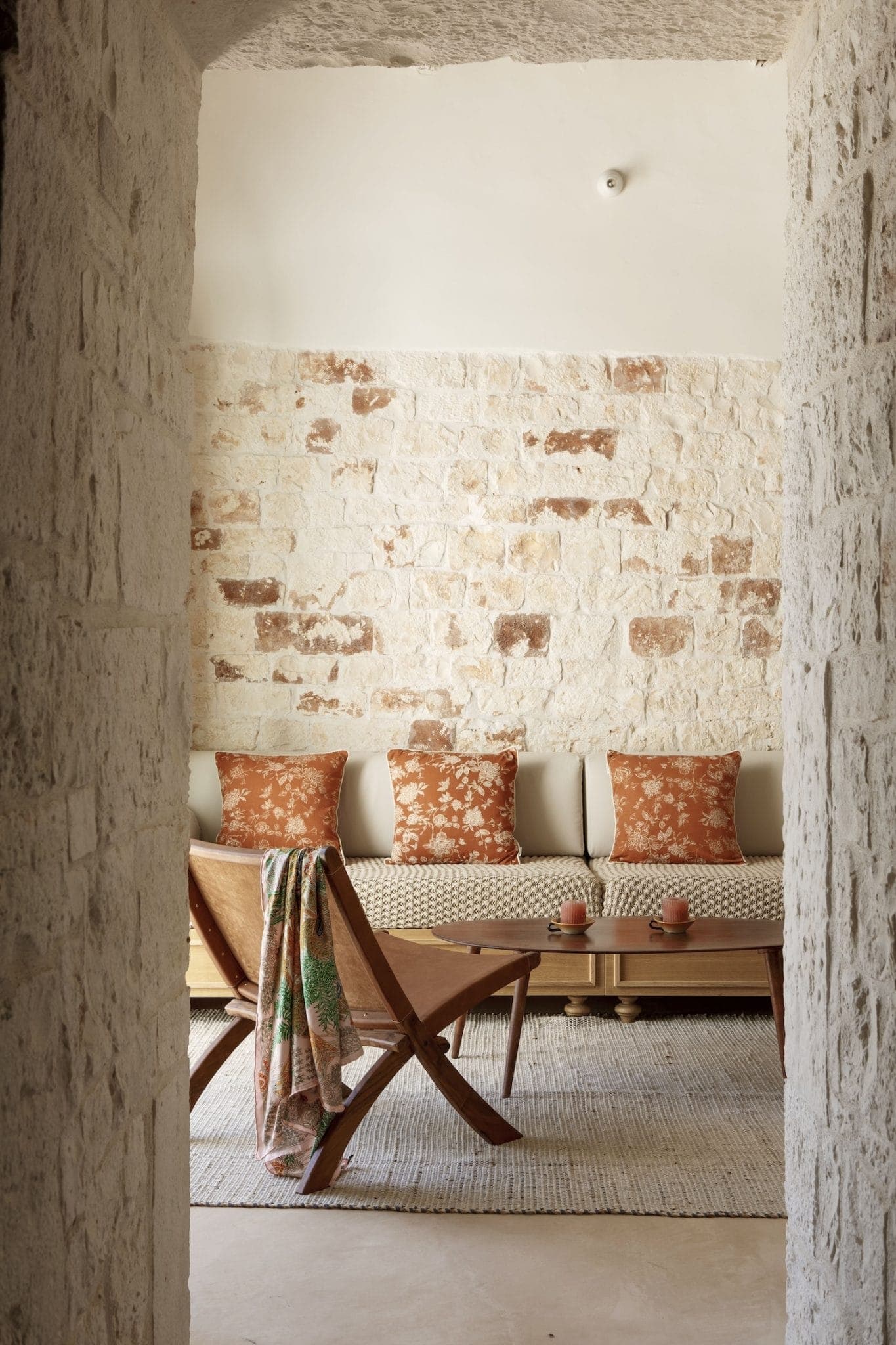 A cozy living room with a stone wall in the background includes a wooden chair and a long sofa adorned with orange floral cushions. A woven rug covers the floor, complementing the warm, earthy tones. A colorful blanket is draped over the chair.