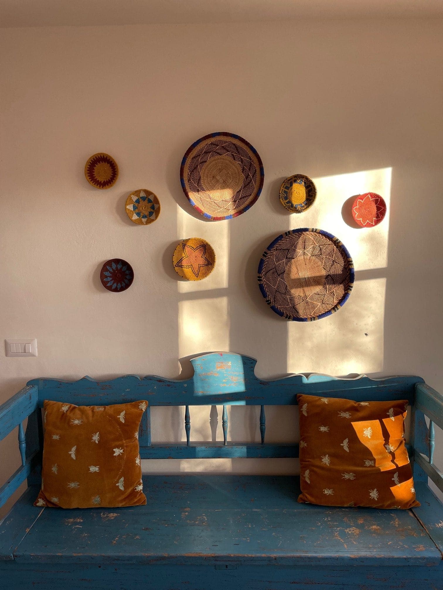 A rustic setting with a blue weathered wooden bench and two orange cushions. Above the bench, various decorative woven baskets and plates are arranged on a sunny white wall, casting soft shadows.