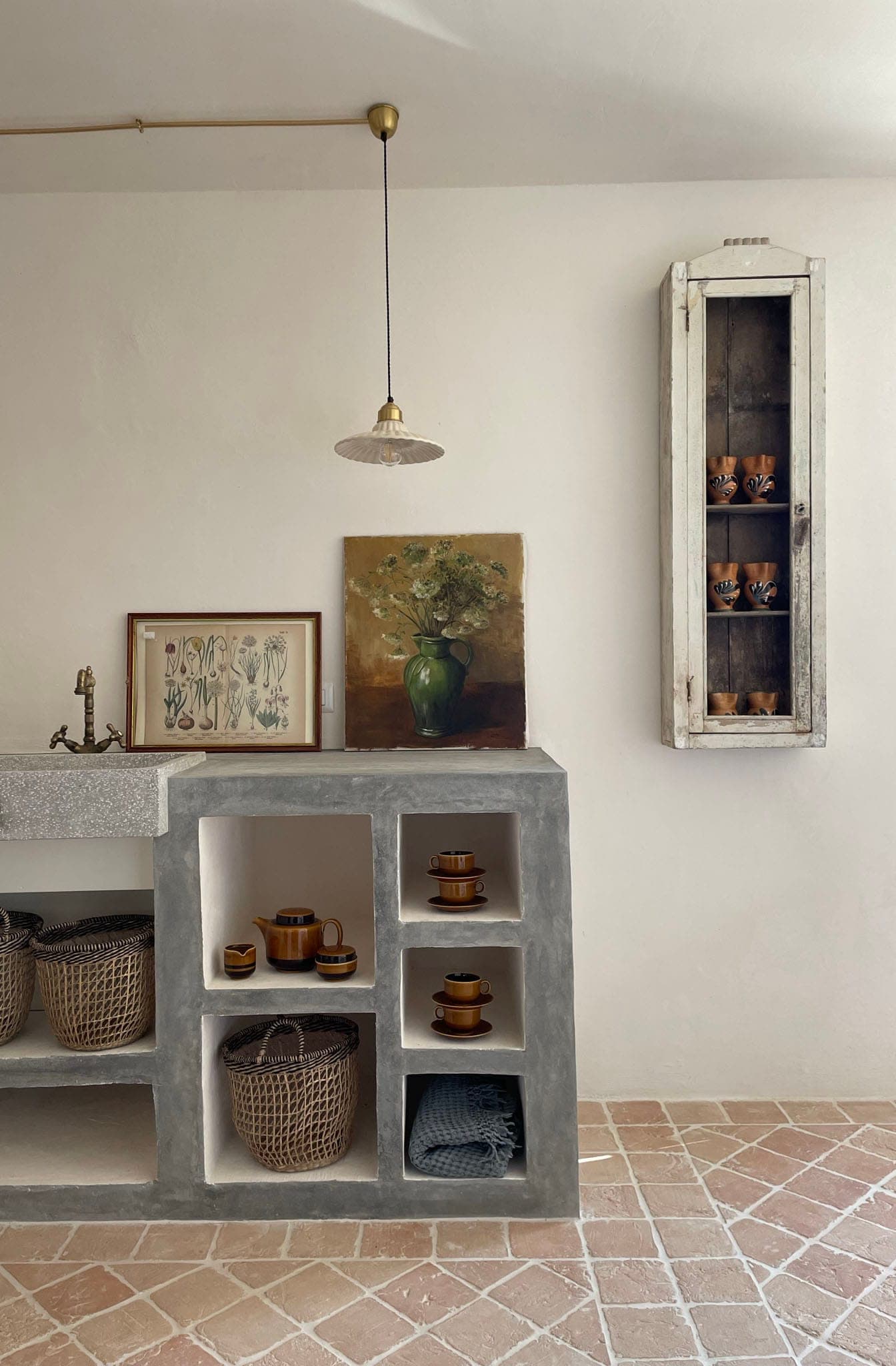 A minimalist kitchen features a stone worktop with open shelving displaying woven baskets and pottery. A vintage painting and botanical print rest against a cream-colored wall, with a hanging lamp above. A narrow cabinet holds more pottery. The tiled floor completes the look.