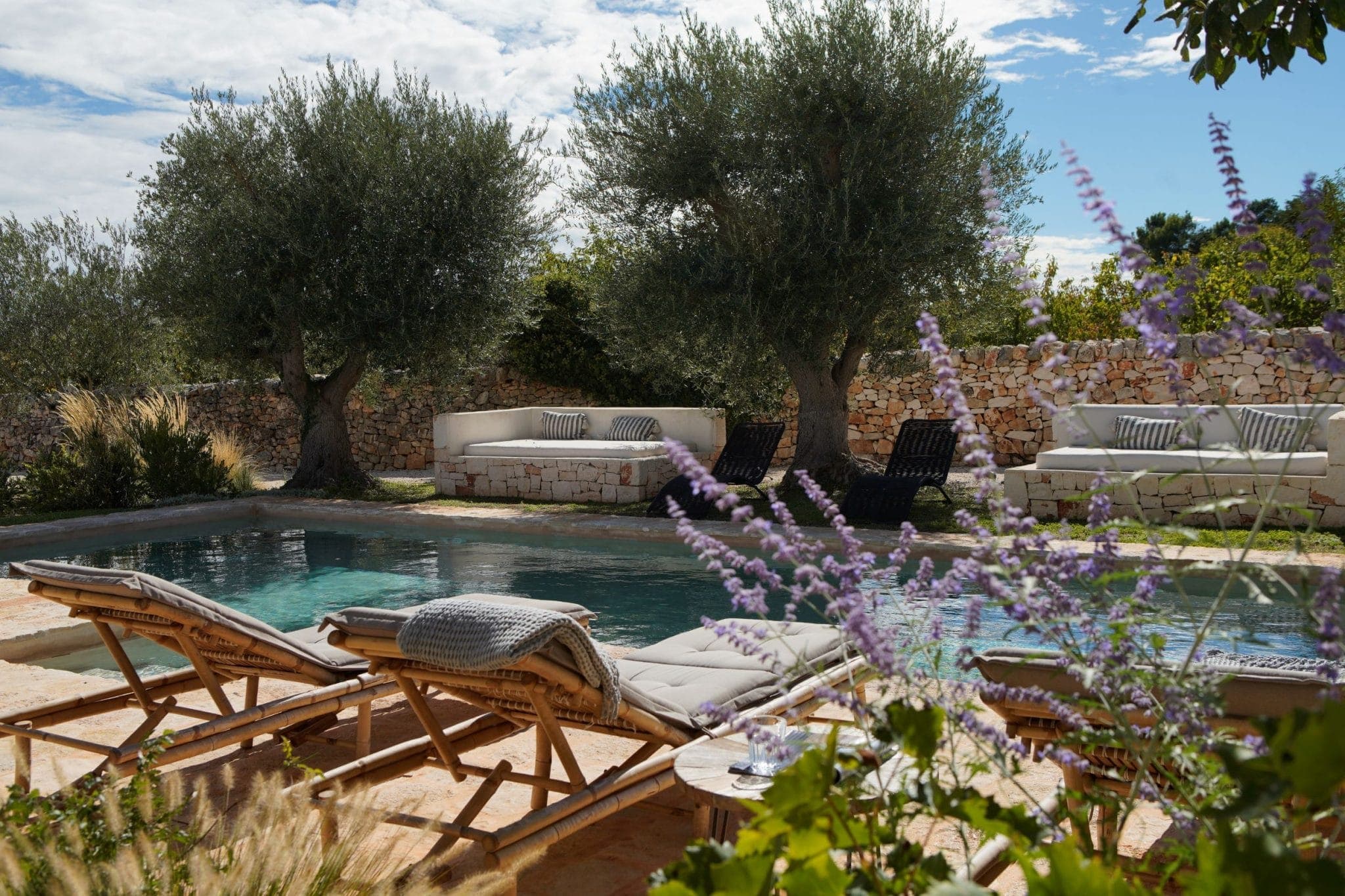 A serene outdoor pool area with loungers facing the water. Lavender flowers line the foreground, while olive trees and stone walls frame the background. Cushioned seating adds to the tranquil ambience under a partly cloudy sky.