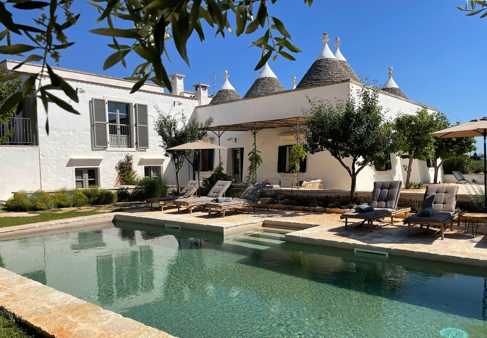 A tranquil outdoor scene includes a swimming pool surrounded by a stone patio and sun loungers. The rustic house in the background has cone-shaped roofs and is shaded by trees. The clear blue sky reinforces the peaceful, summery atmosphere.