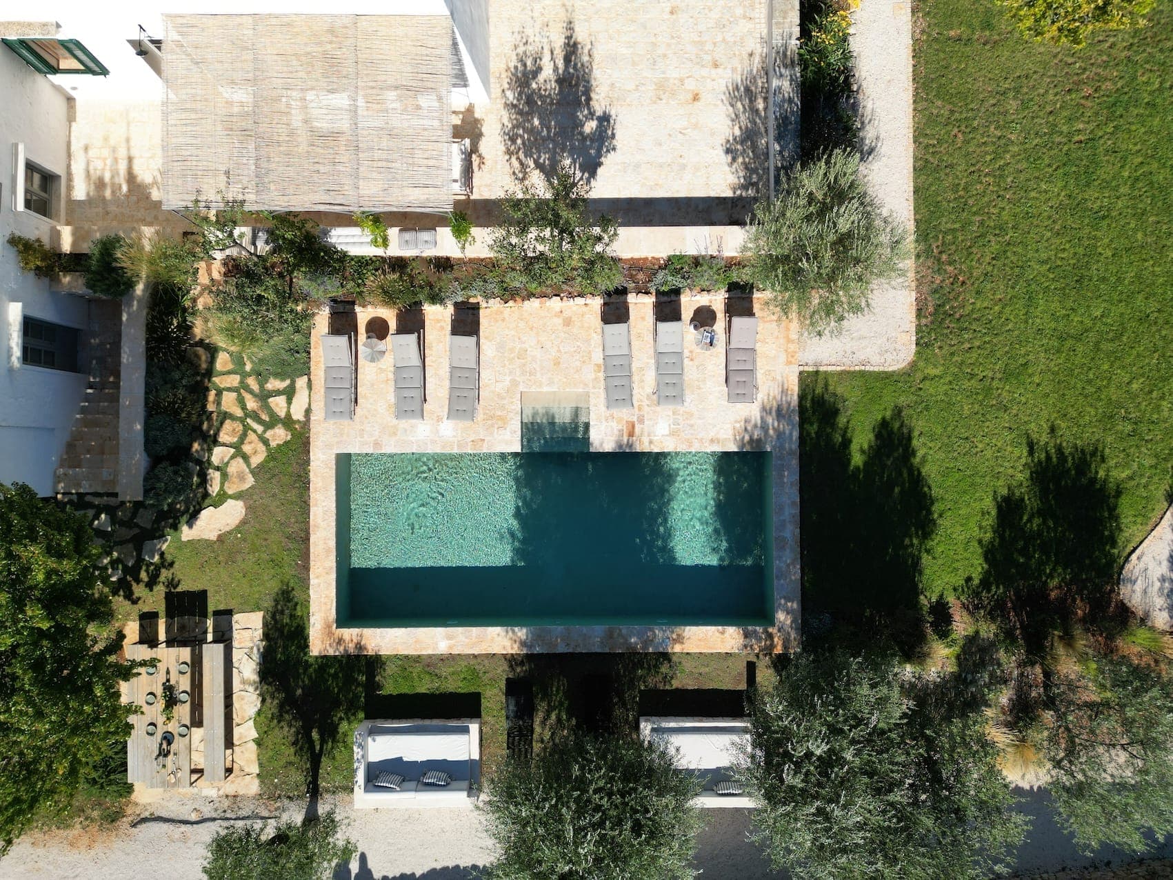 Aerial view of a rectangular pool surrounded by sun loungers and deckchairs on a stone patio. The pool is adjacent to a thatched-roof building. The area is bordered by green grass and trees. A garden can be seen nearby.