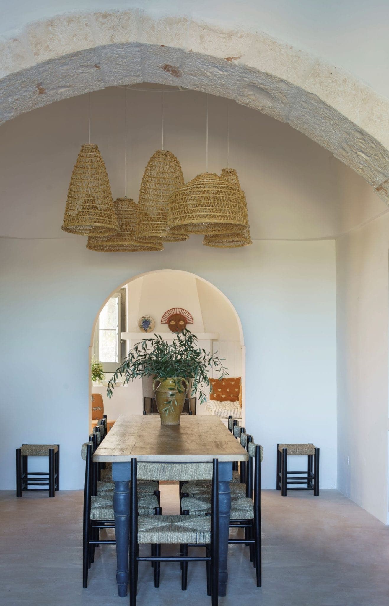A rustic dining room with a long wooden table surrounded by wicker chairs. Above, several woven basket-style light fixtures hang. An archway leads to a room with plants and orange cushions visible in the background.