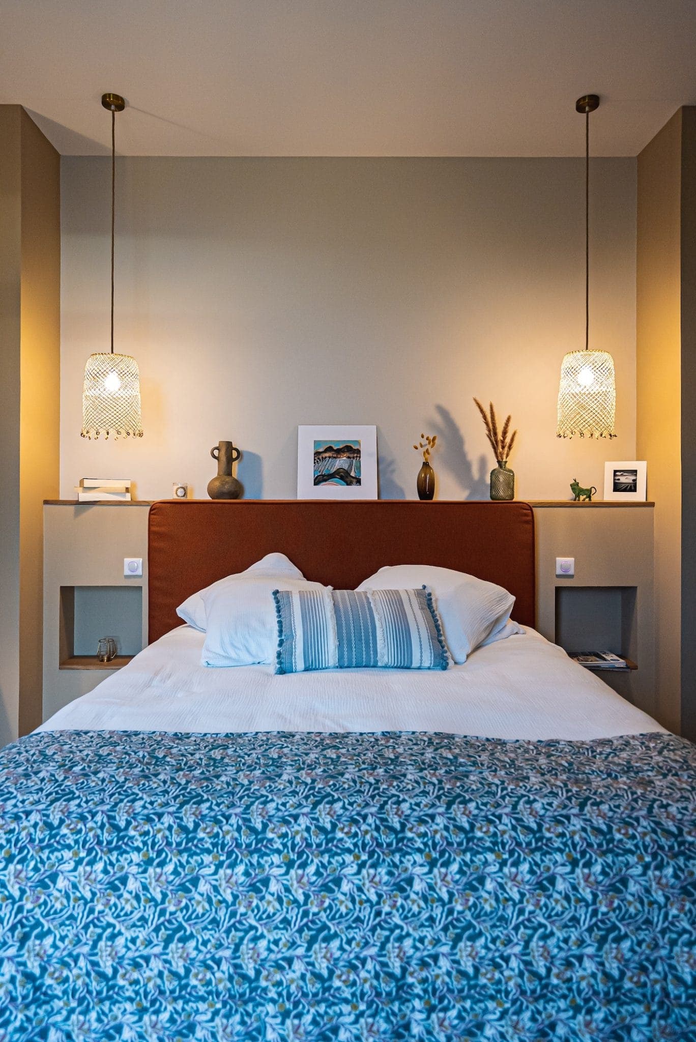 A comfortable room with a double bed with a patterned blue blanket and white pillows. Two hanging lamps are suspended above the bed and the wall shelf features decorations including framed artwork and vases. The atmosphere is warm and relaxing.