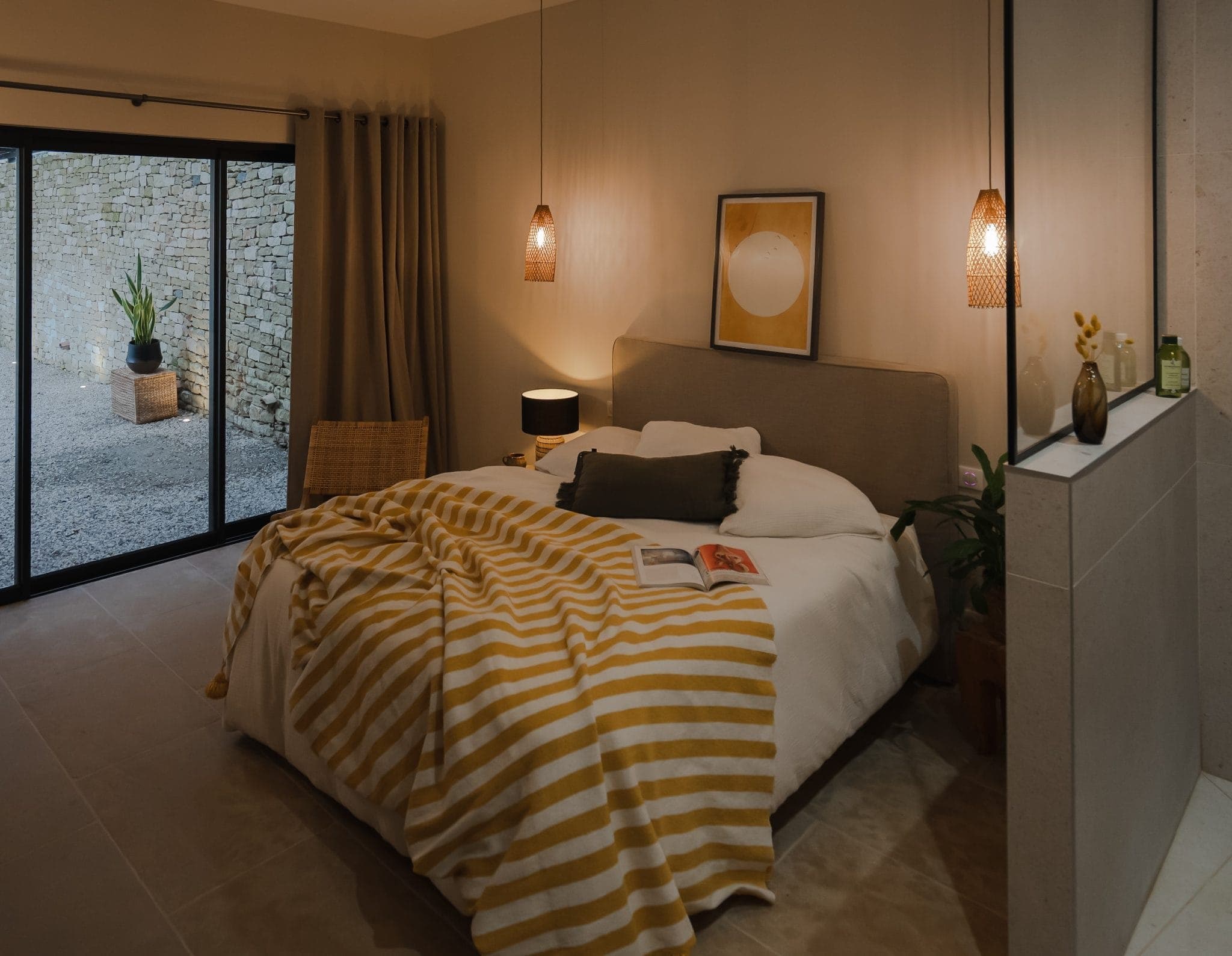 A cosy bedroom with a large bed covered with a yellow and white striped blanket. Two hanging lamps are suspended above the bed. A sliding glass door opens onto an outdoor space with plants. Wall art and a small table lamp complete the decor.