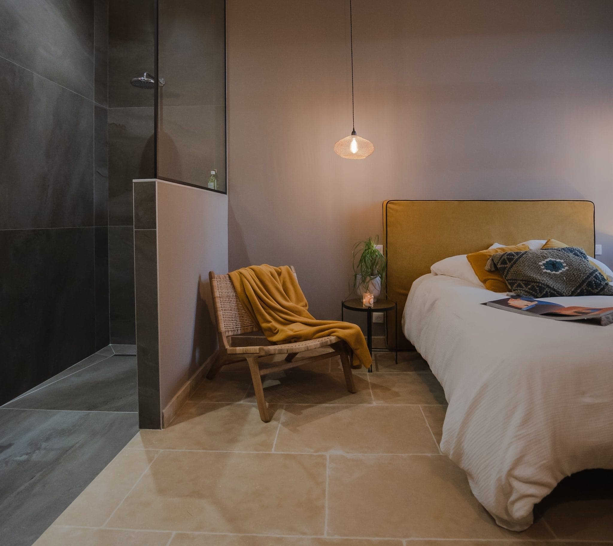 A comfortable bedroom includes a bed with mustard-yellow accents, a side table with a plant, a hanging lamp and a wicker chair with a mustard plaid. An open shower with dark tiles sits in the corner, complementing the neutral tiled floor.